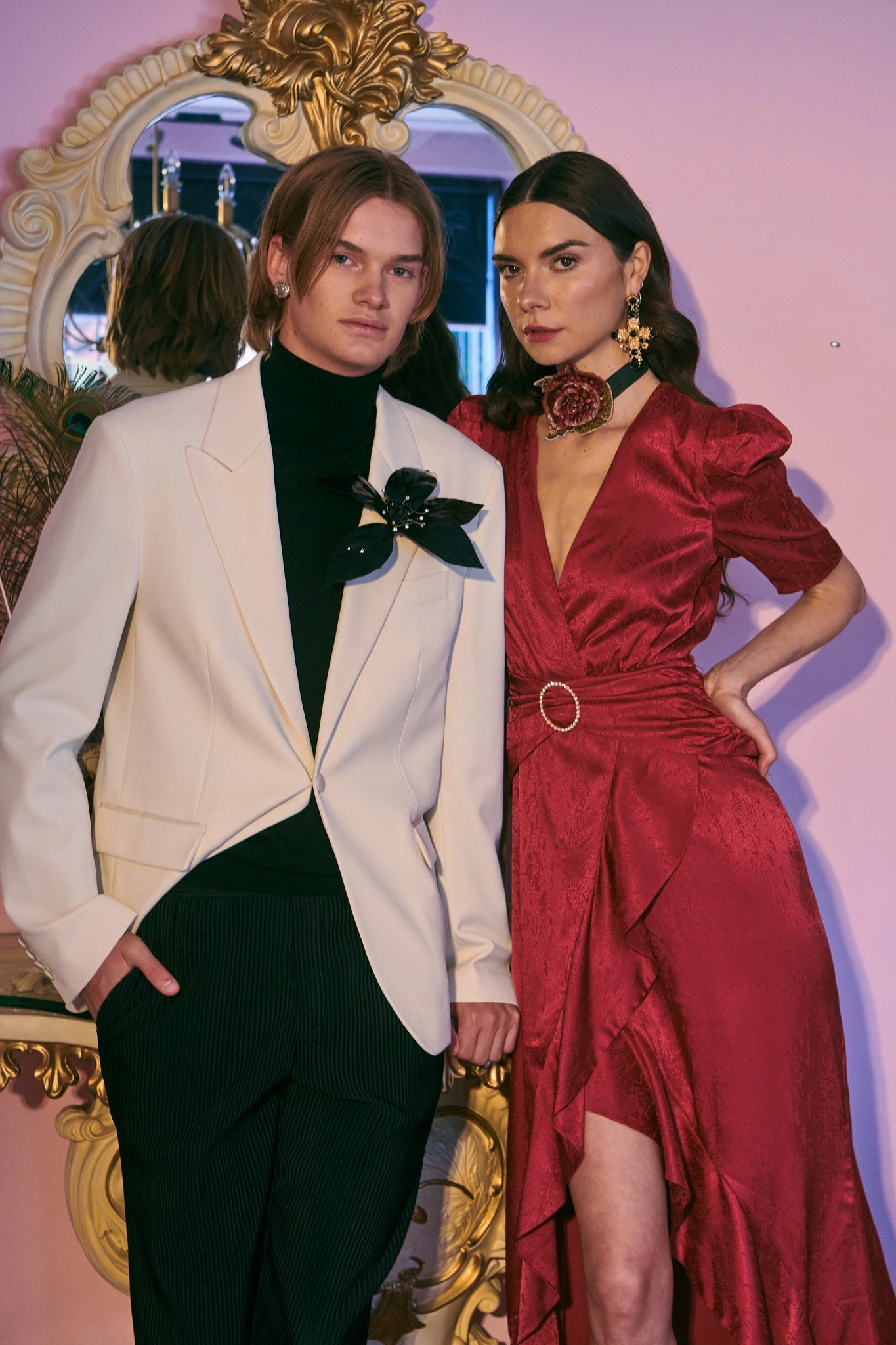 Two people posing in formal attire against a decorative mirror. There is a man in a white suit jacket. He is wearing a broochella giglio flower brooch. The lady is dressed in red and has dark hair.