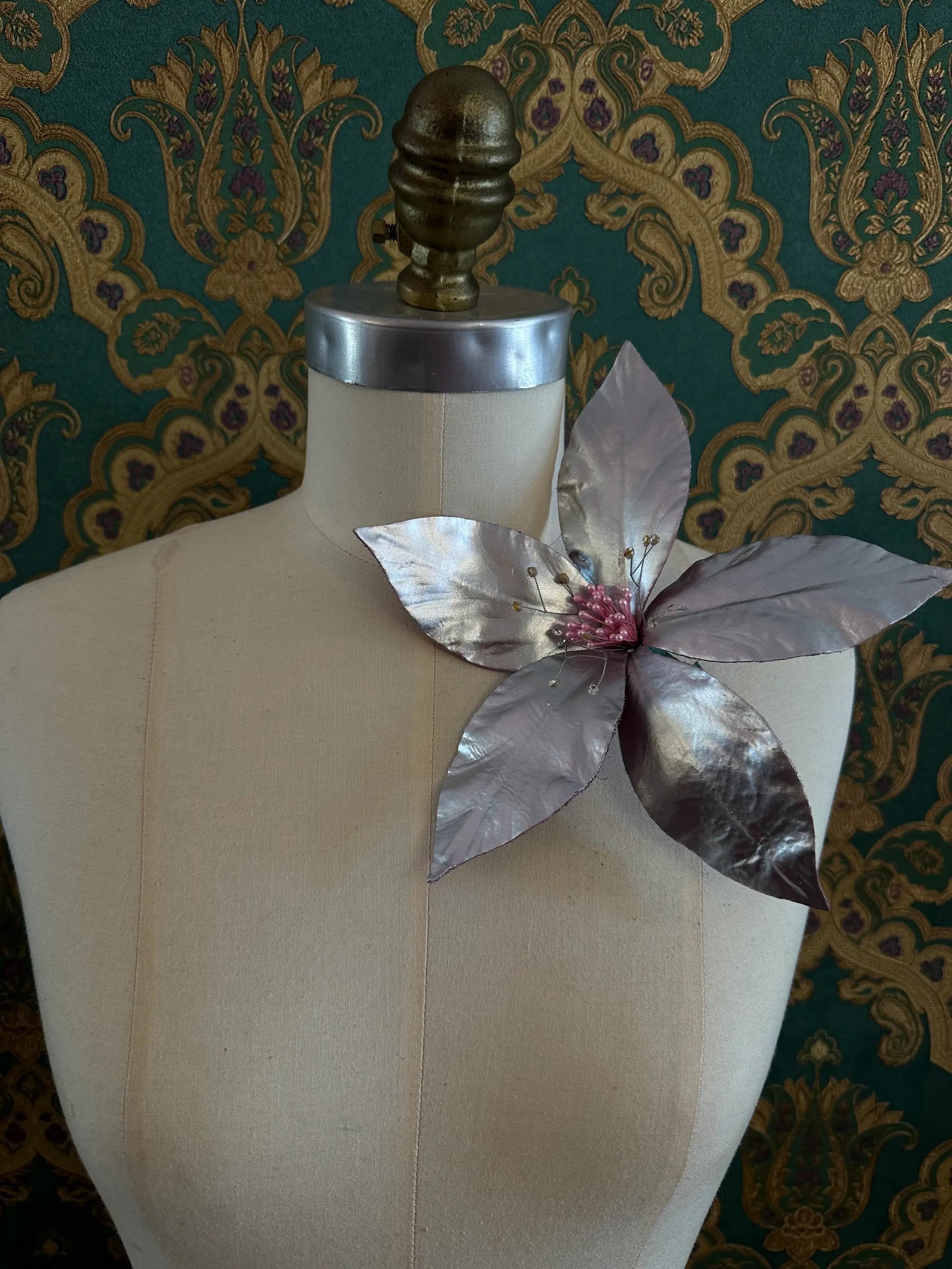 Broochella Giglio silver patent leather large lily flower brooch. Handmade in New York. It is presented on a mannequin. The background is green and gold damask wallpaper.