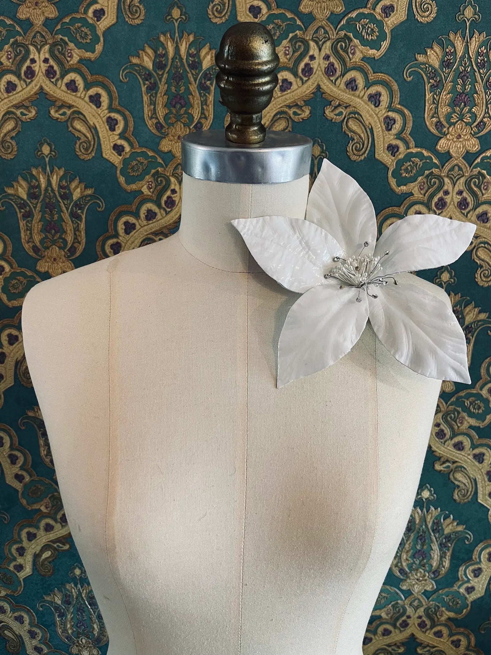 Broochella giglio handmade large patent leather flower in white. Styled on a mannequin. The background is green and gold damask wallpaper