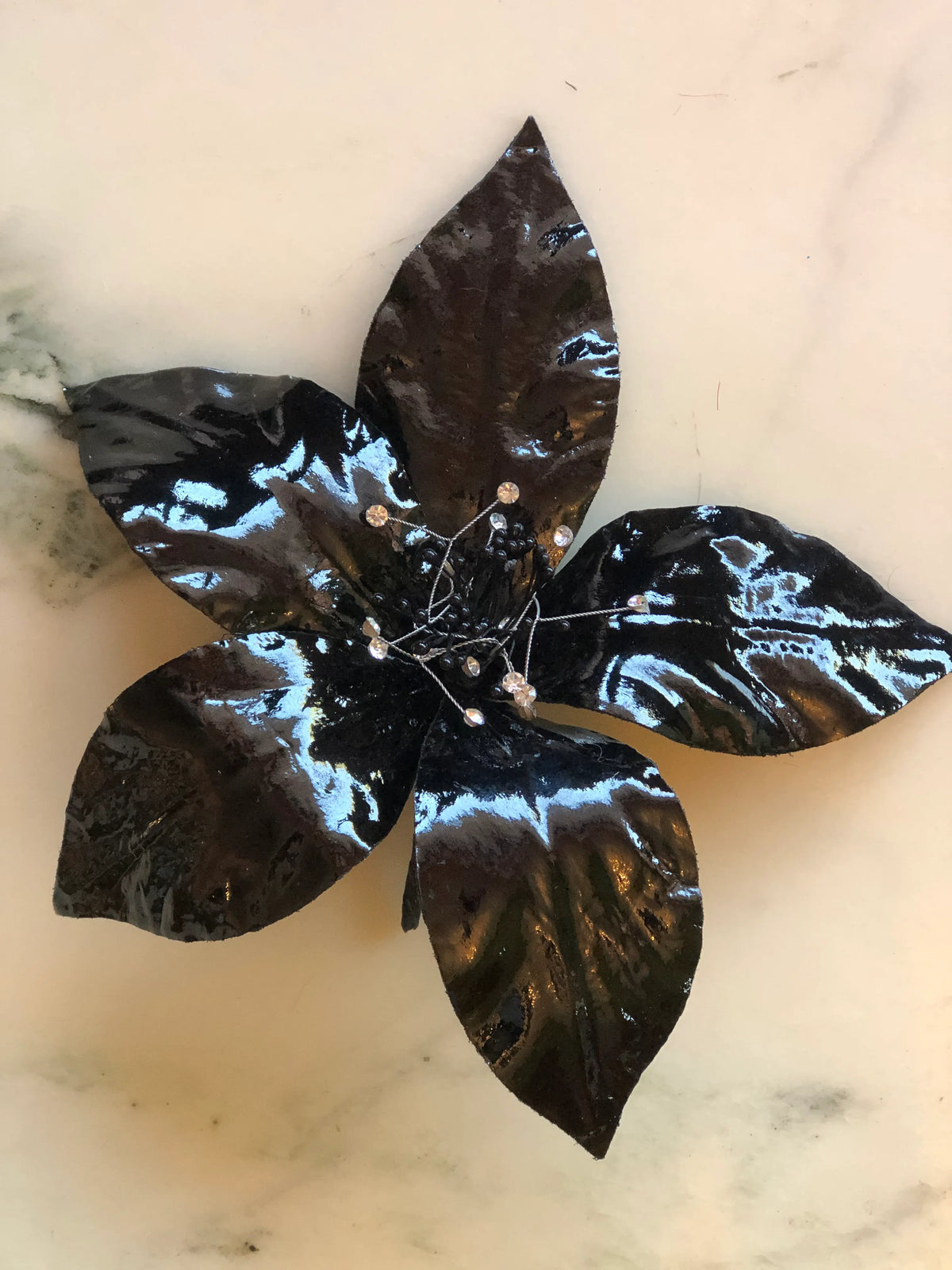 Broochella Giglio Flower Brooch Pin in glossy black, featuring sculptural leather lily petals with a textured black centre and delicate crystal-tipped stamens on a cream marble background.