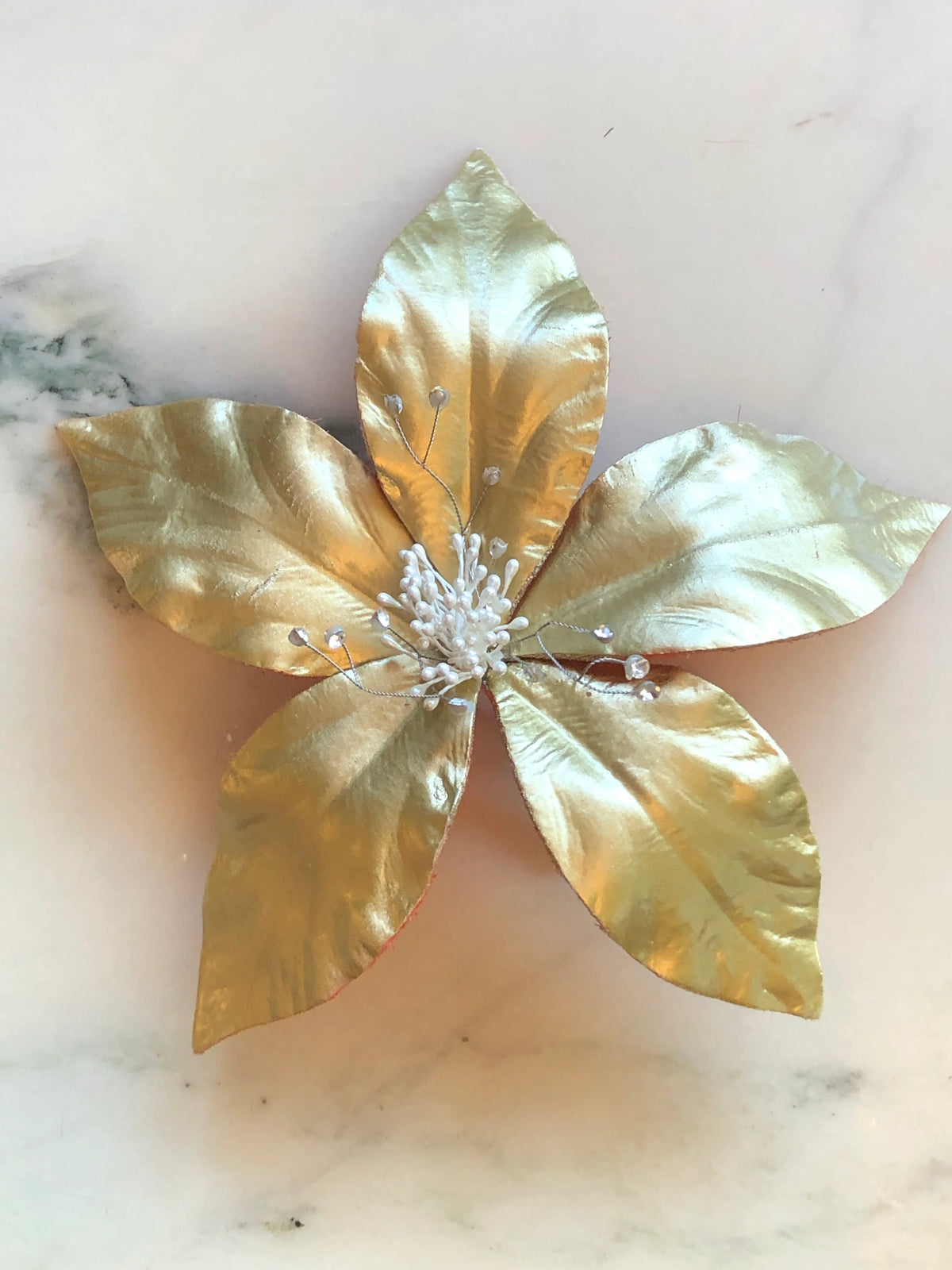 Broochella Giglio Flower Brooch Pin in glossy gold featuring sculptural leather lily petals with a textured black centre and delicate crystal-tipped stamens on a cream marble background.