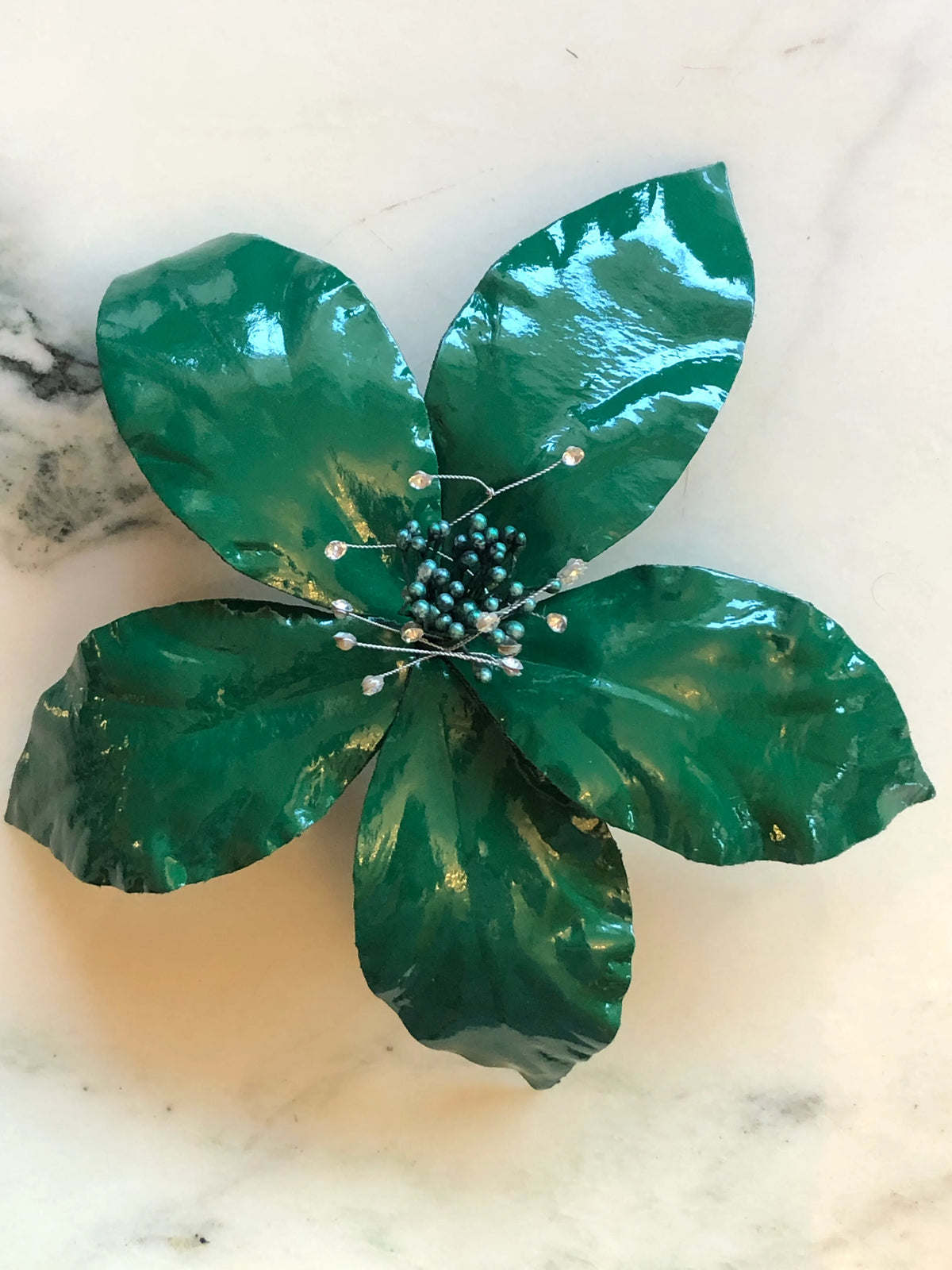 Broochella Giglio Flower Brooch Pin in glossy green, featuring sculptural leather lily petals with a textured black centre and delicate crystal-tipped stamens on a cream marble background.