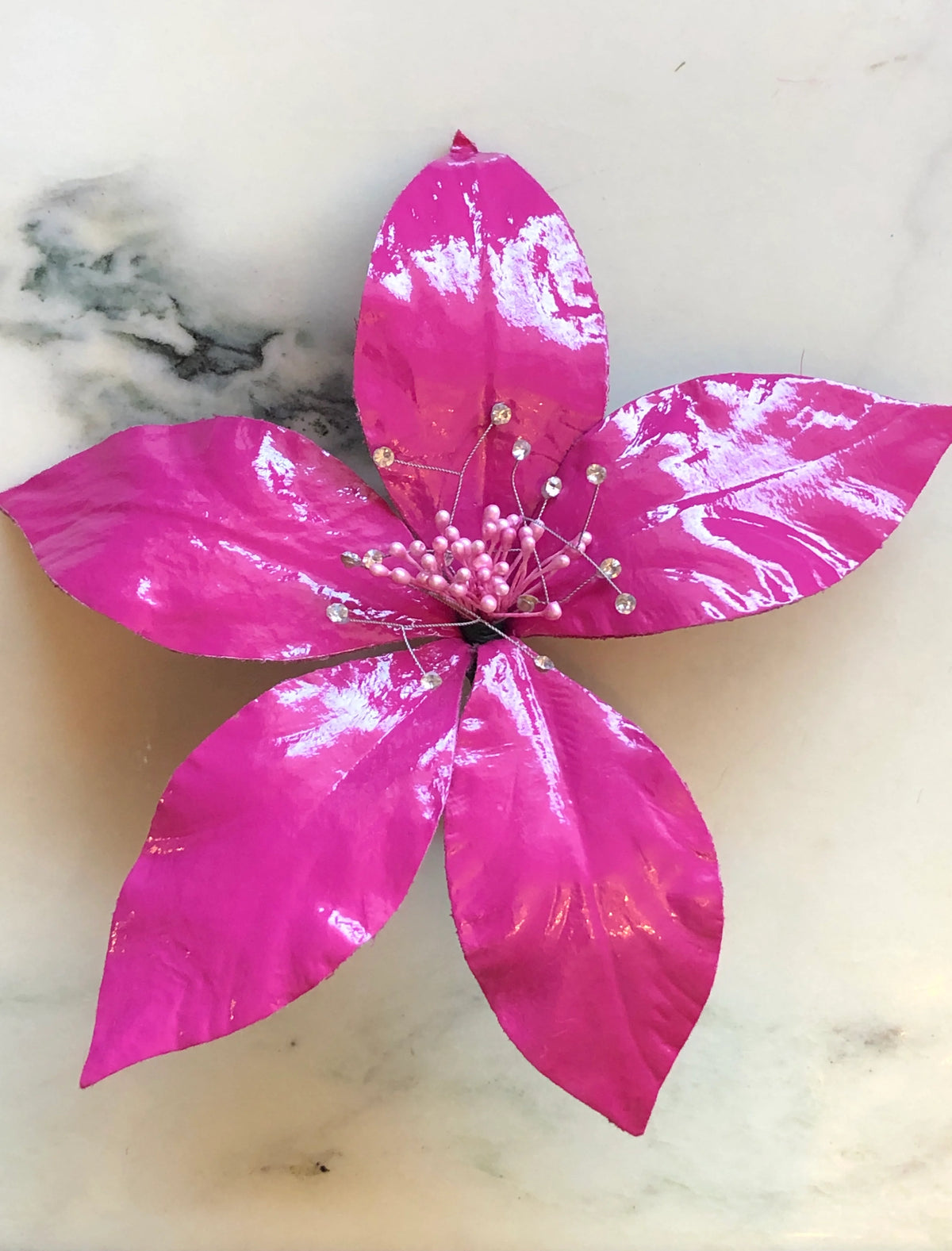 Broochella Giglio Flower Brooch Pin in glossy pink, featuring sculptural leather lily petals with a textured black centre and delicate crystal-tipped stamens on a cream marble background.