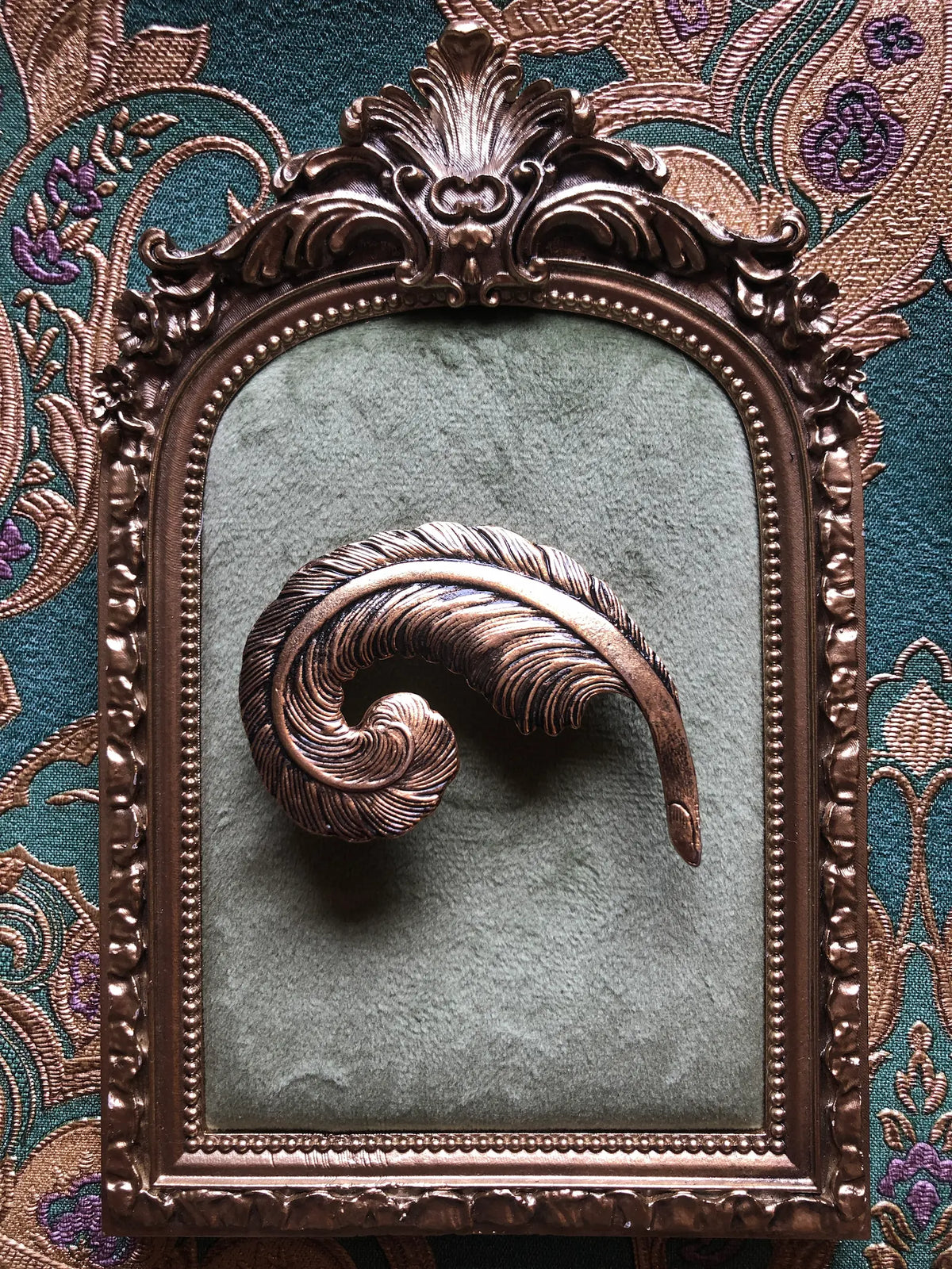 Broochella Gilded Plume vintage feather brooch with an antiqued gold-tone finish, curled sculptural shape and deeply engraved detailing, measuring approximately 5 × 7 cm. Photographed on a green velvet tray with an ornate gold frame. The background is green and gold damask wallpaper.