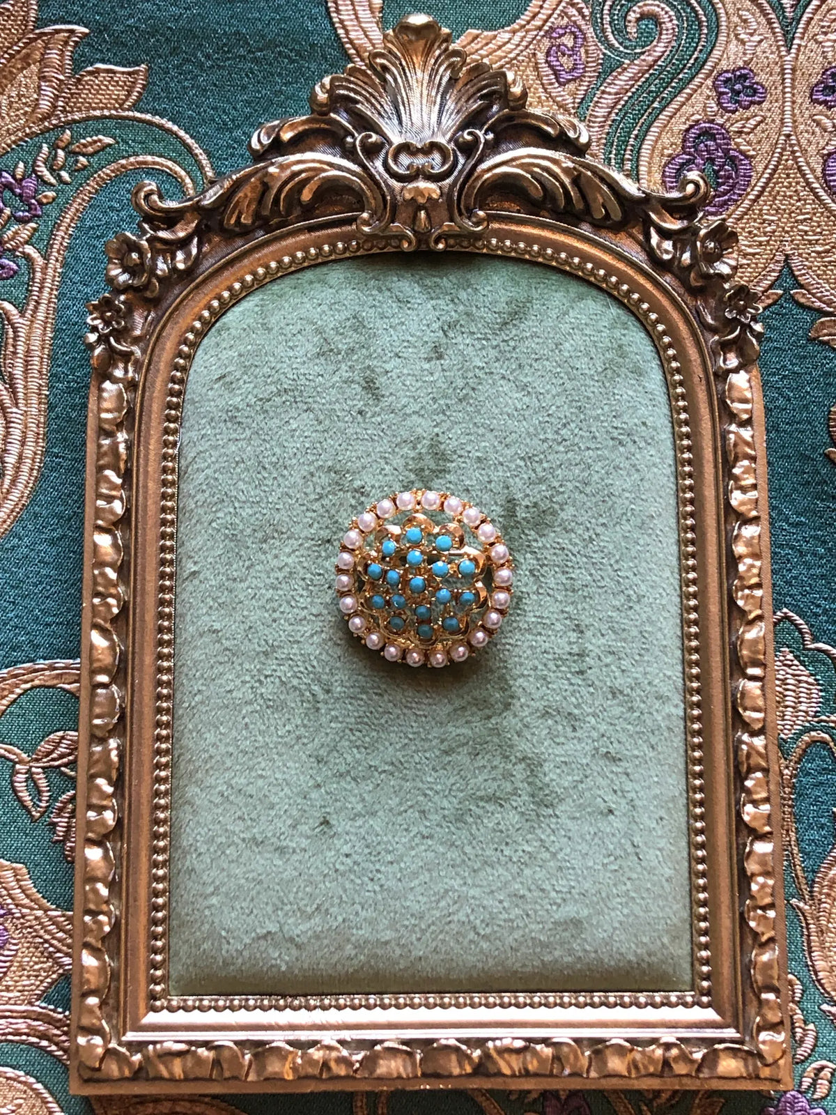 Broochella Ginevra Vintage Turquoise & Pearl Brooch featuring turquoise-blue cabochons, faux pearls and ornate gold-tone openwork. Pictured on a green velvet tray with an ornate gold frame. The background is green and gold damask wallpaper.