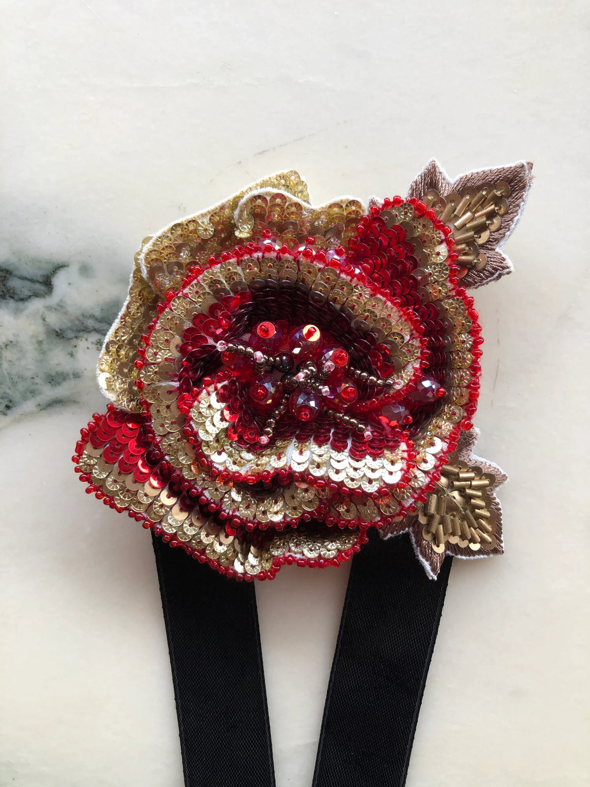 Broochella Girocollo Bejewelled Flower Choker & Brooch Pin featuring a sculptural red and gold sequinned flower with beaded detailing, metallic leaves and long black ribbon ties, photographed on a white marble background with soft grey-green veining.