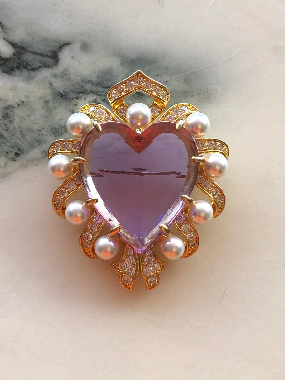 Broochella Giulietta e Romeo Brooch Pin featuring a large faceted lilac heart crystal framed by white pearls and sparkling clear crystals in a gold-tone setting. Pictured on a white marble surface