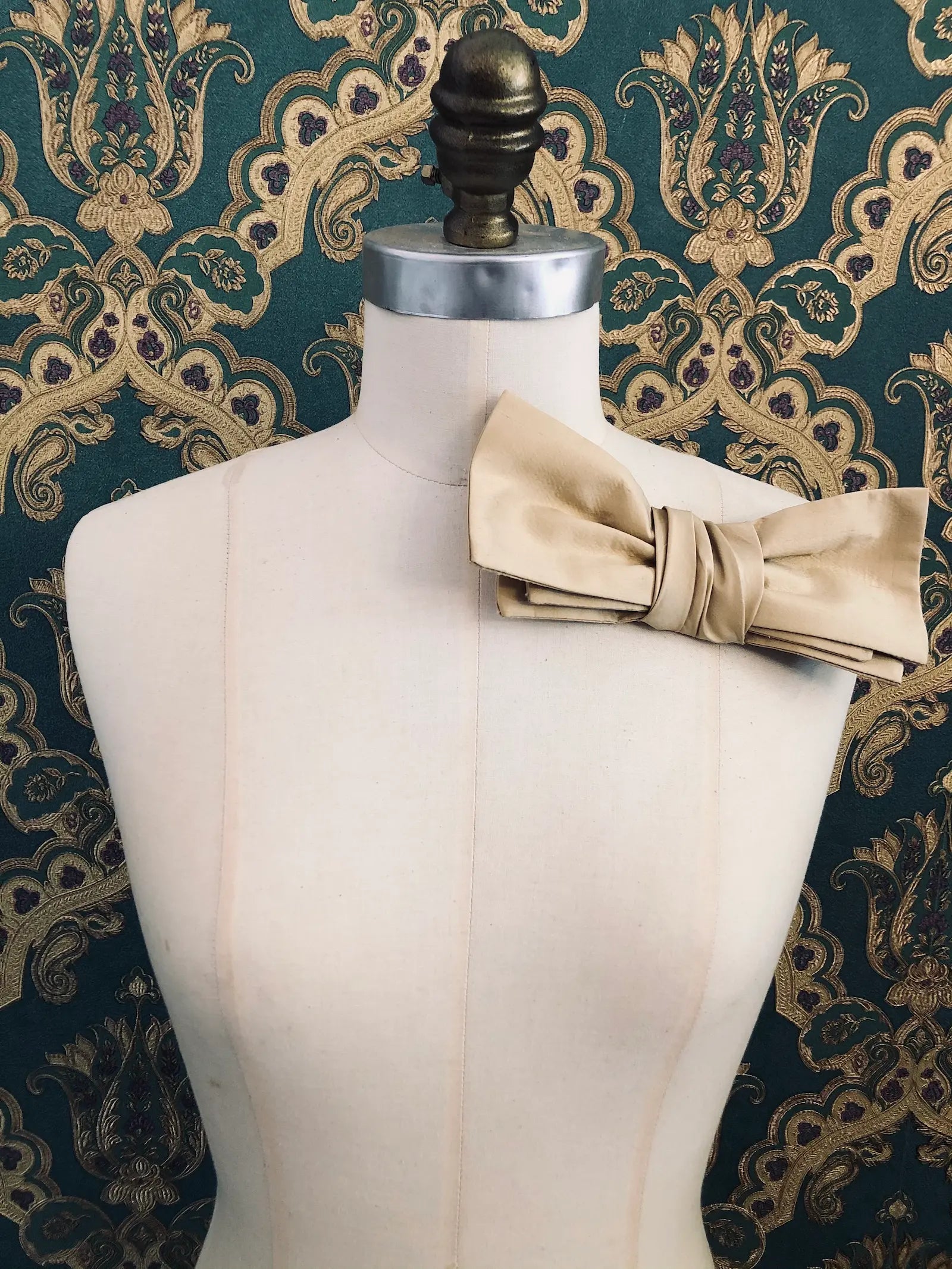 Broochella Marie Antoinette handmade, designer silk bow brooch in gold. It is pinned to a mannequin. The background is green and gold damask wallpaper.