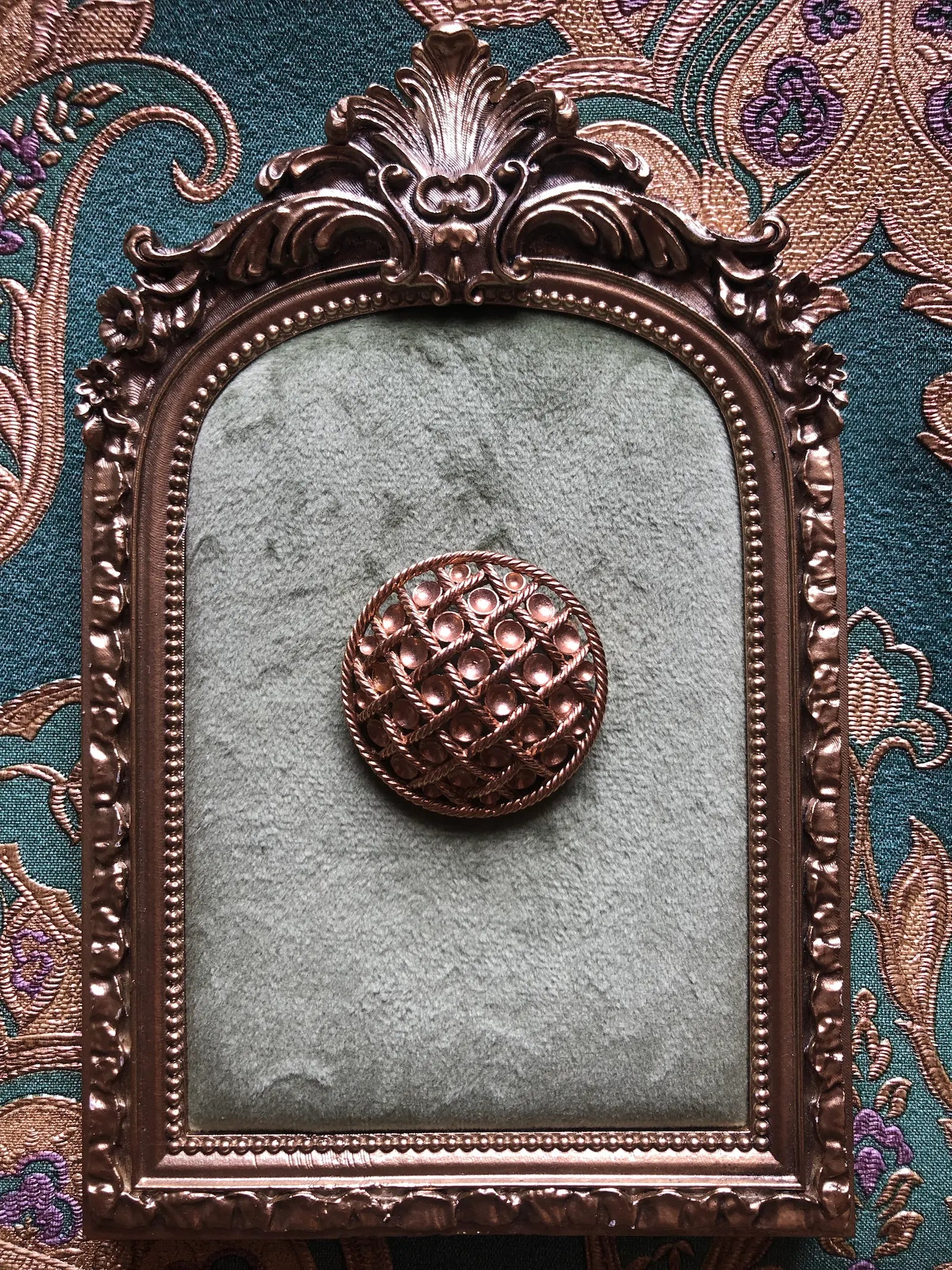 Broochella Golden Constellation vintage round brooch with raised gold-tone discs and an intricate twisted rope lattice, measuring approximately 4 cm across. Photographed on a green velvet tray with an ornate gold frame. The background is green and gold damask wallpaper.