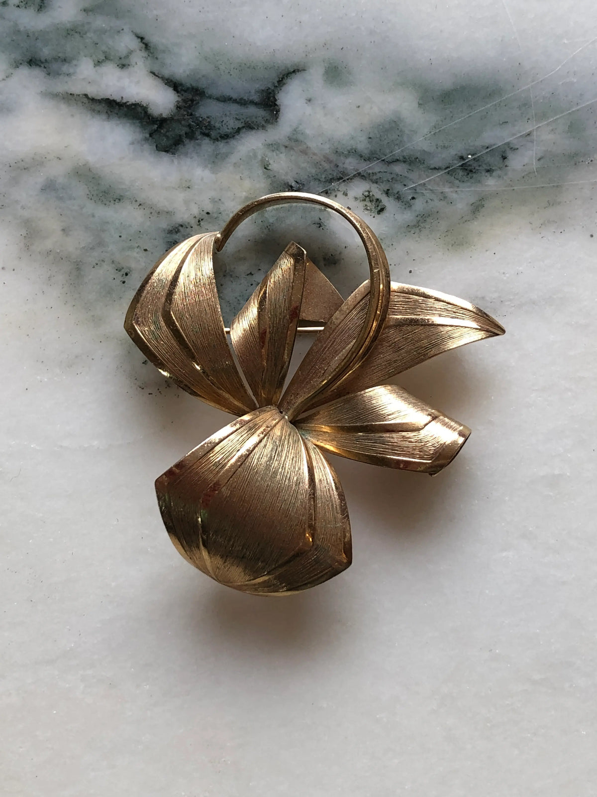 Broochella Golden Flourish vintage gold-tone brooch with an abstract bow, sweeping ribbon shapes and fine textured detailing, measuring approximately 6 × 5 cm. Photographed on white and grey marble.