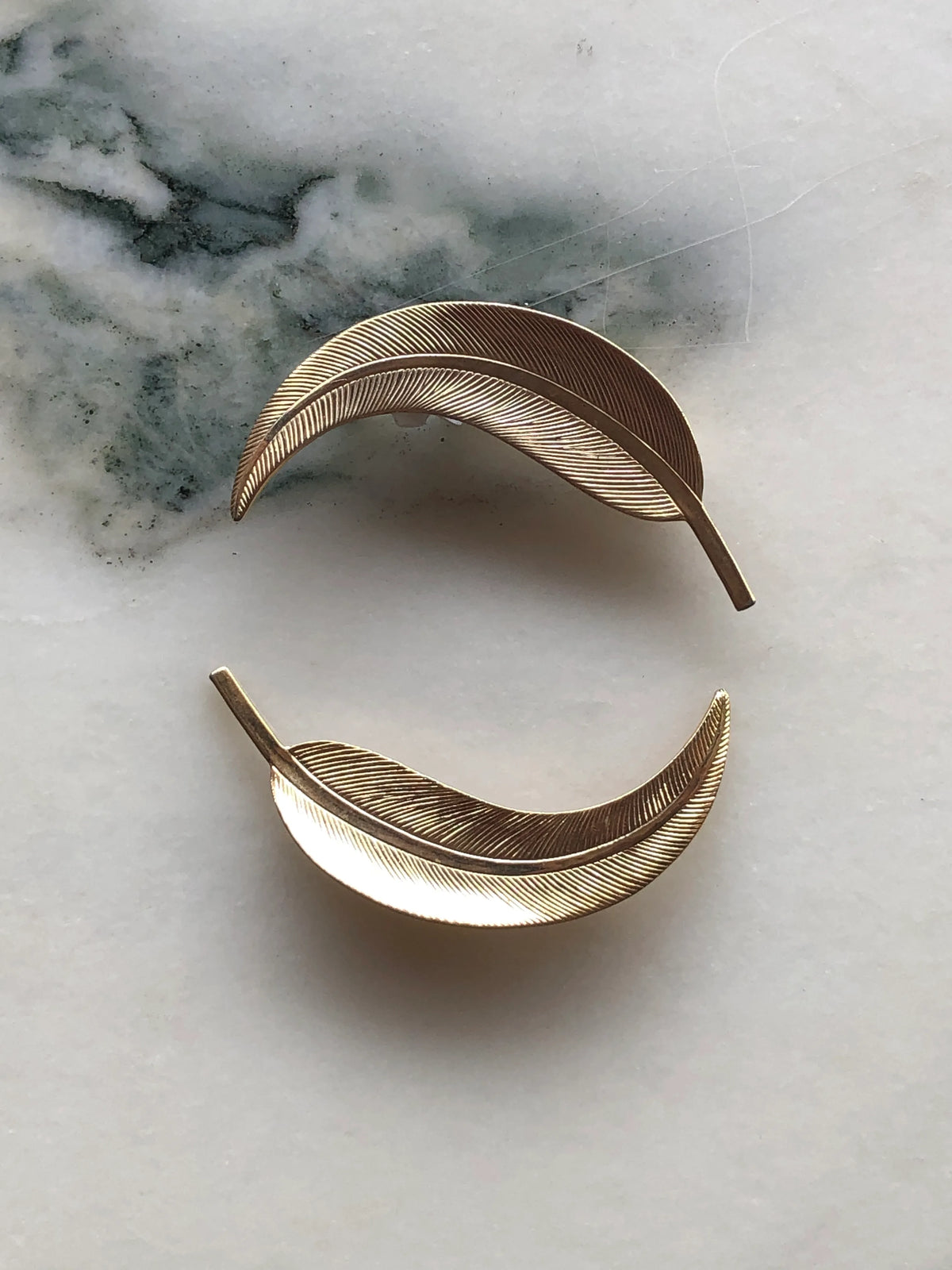 Broochella Golden Gum Leaf Vintage Brooch Pair featuring two sculptural curved gold-tone leaves with finely etched vein detailing, photographed on a white marble background with soft grey-green veining.