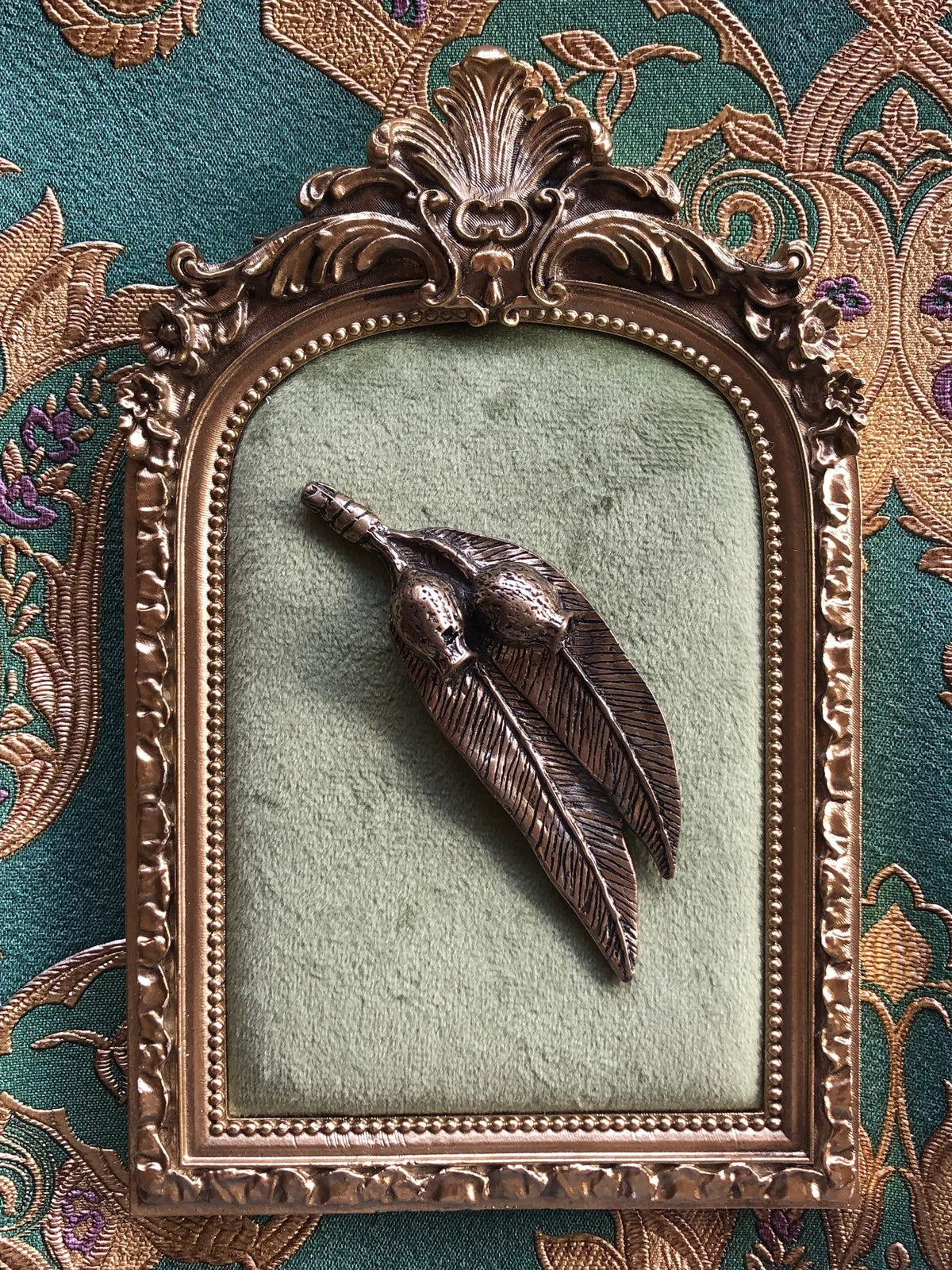 Broochella Golden Gumnut Trail Vintage Brooch featuring two sculpted Australian gumnuts and long eucalyptus leaves in antiqued bronze-tone metal, displayed on sage green velvet in an ornate gold frame against green and gold patterned fabric.