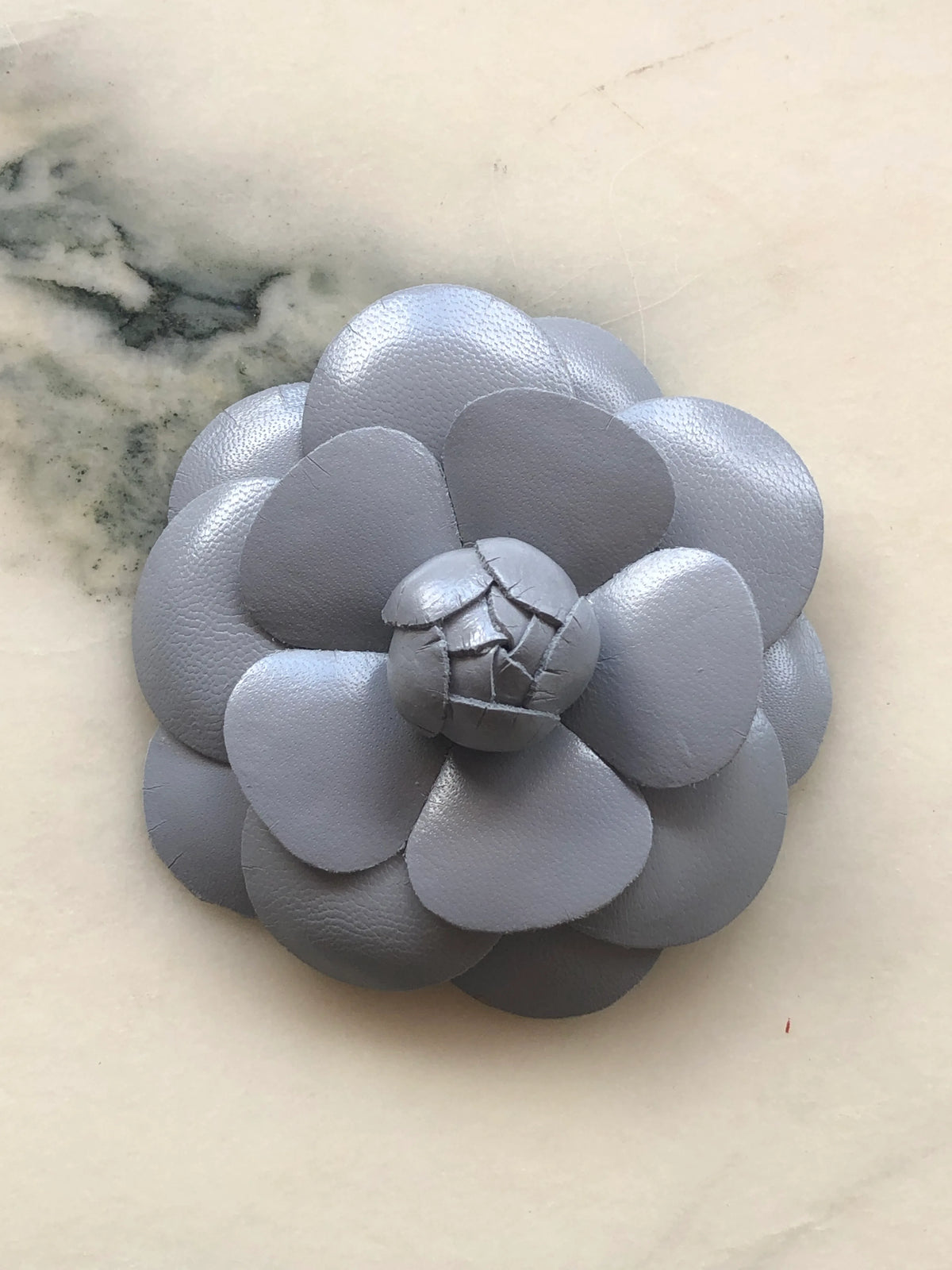 A Broochella Camelia handmade black leather flower-shaped accessory brooch,  resting on a white marble surface.