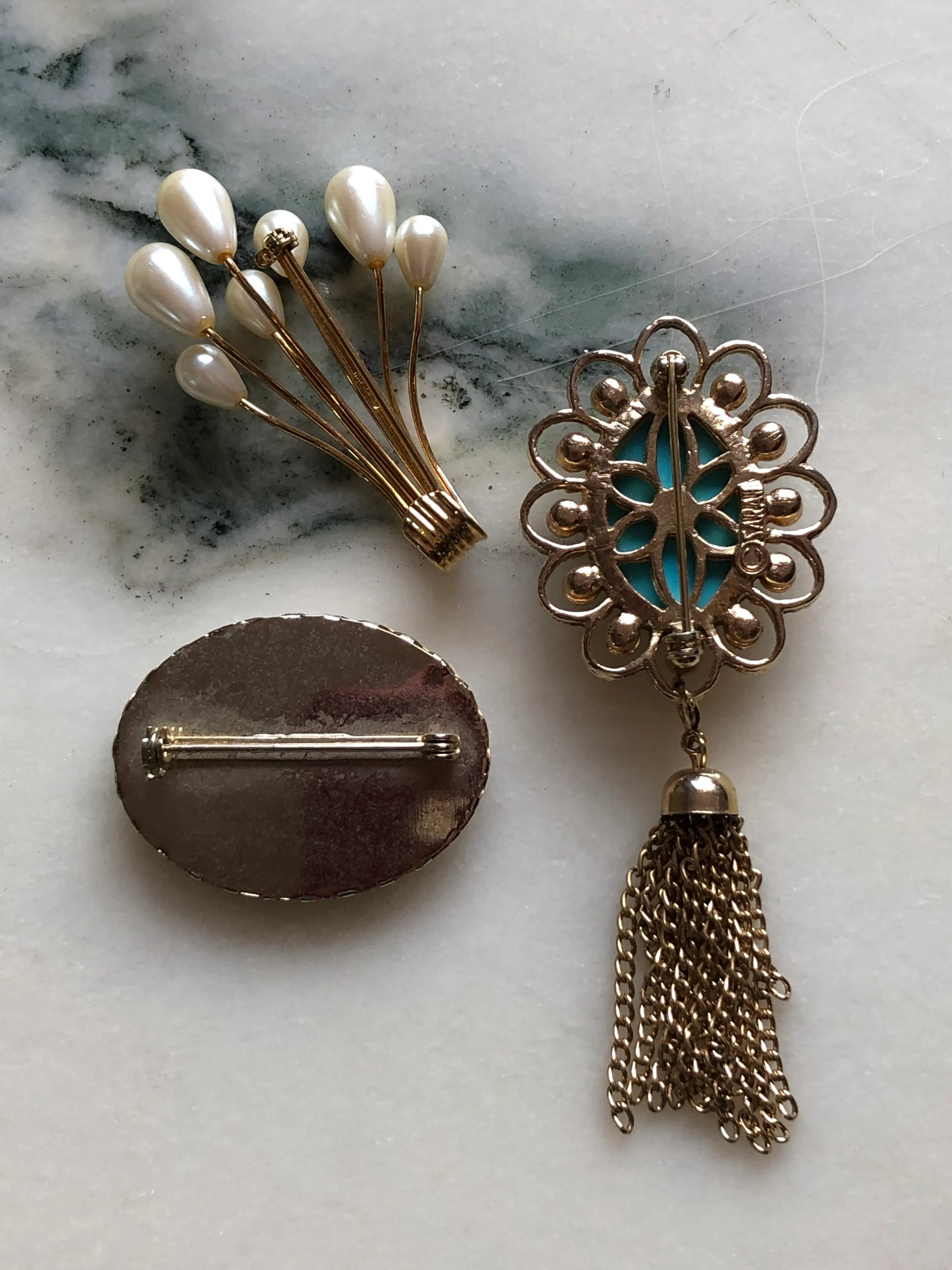Broochella The Heirloom Trio vintage brooch set featuring a gold-tone faux-pearl spray, an oval floral brooch and an ornate turquoise-blue cabochon brooch with faux pearls and a chain tassel. Photographed on white and grey marble.