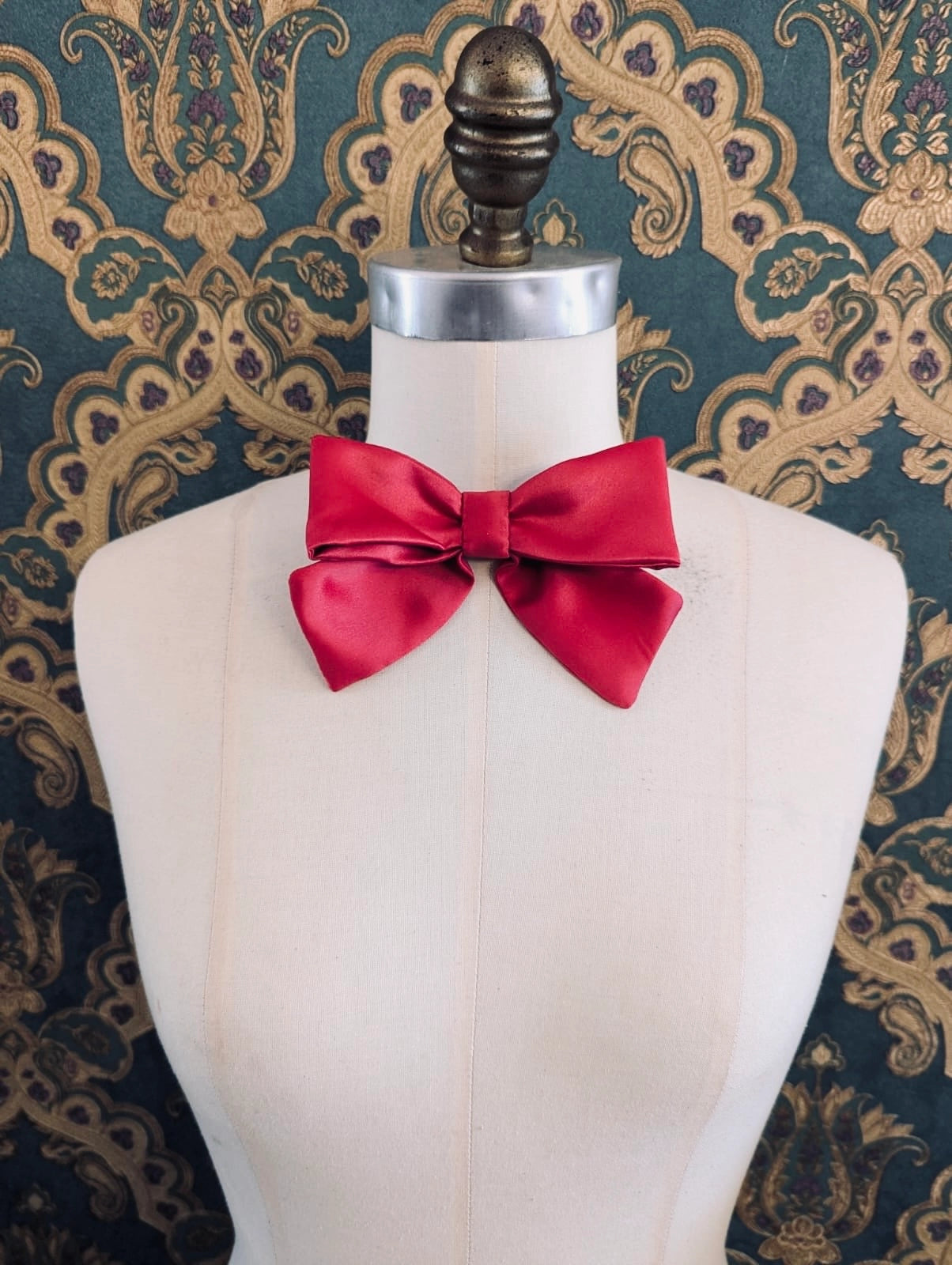 Broochella hollywood handmade satin bow in pink. The brooch is pinned to a mannequin at the neck. The background is green and gold damask wallpaper.