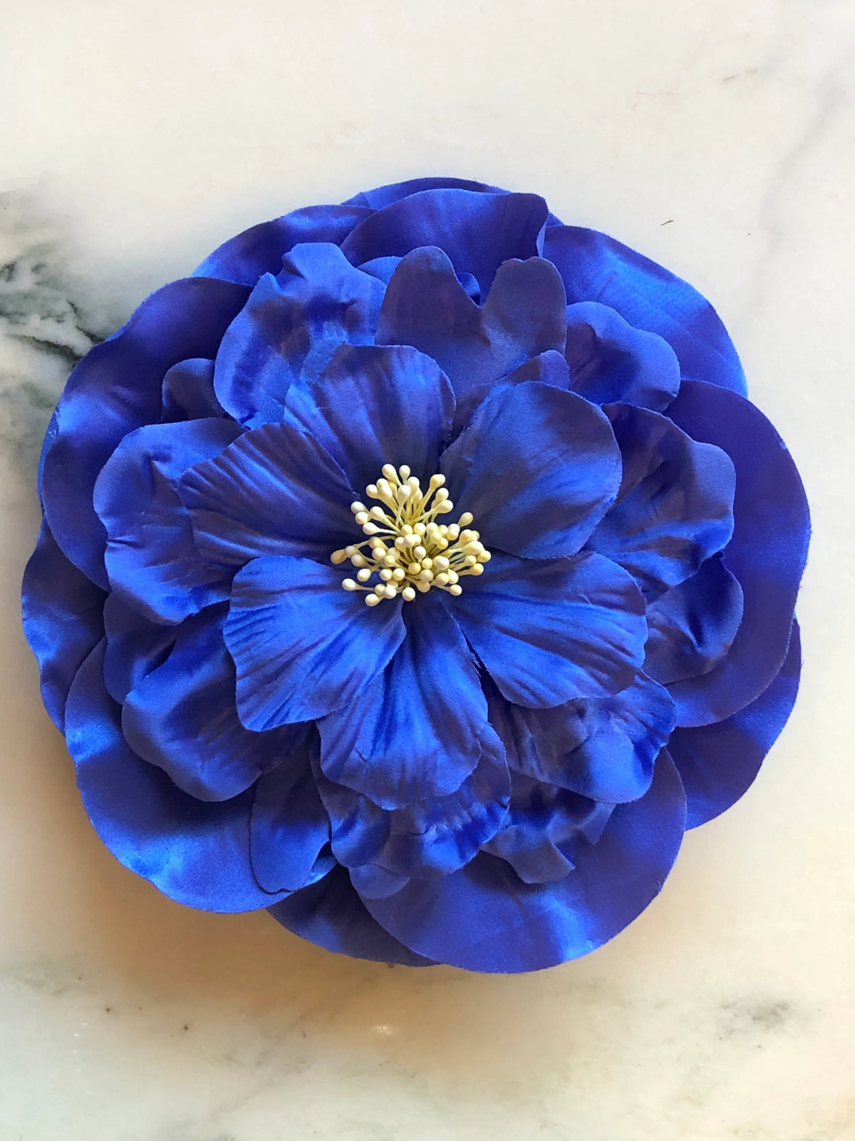 Broochella Ibisco Flower Brooch in vivid cobalt blue, featuring full layered fabric petals with sculptural texture and a contrasting cream stamen centre on a cream marble background.