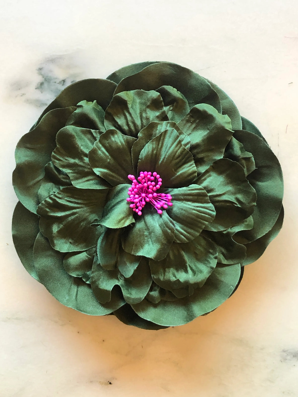 Broochella Ibisco Flower Brooch in rich green, featuring full layered fabric petals with sculptural texture and a contrasting bright pink stamen centre on a cream marble background.