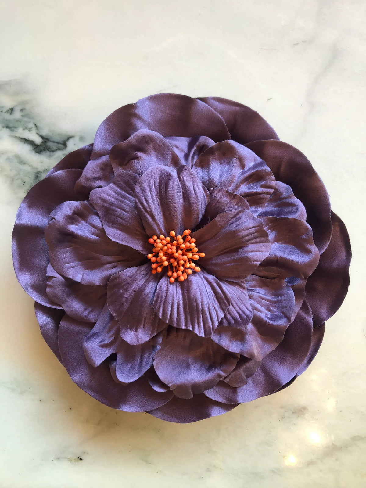 Broochella Men's  Ibisco Flower Brooch in soft purple, featuring full layered fabric petals with sculptural texture and a contrasting orange stamen centre on a cream marble background.