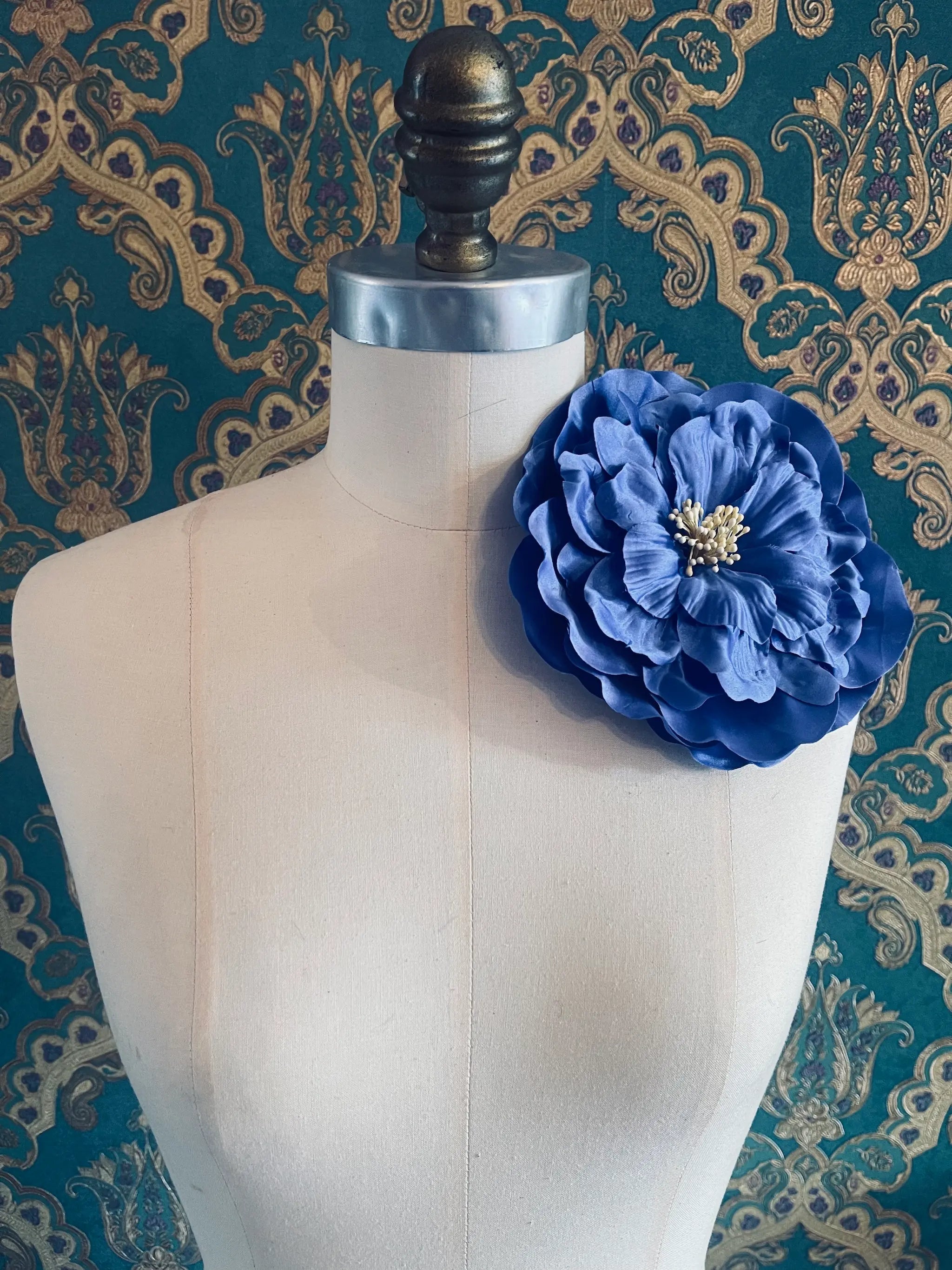 Broochella Ibisco large, blue, handmade flower brooch. Styled on a mannequin. The background is green and gold damask wallpaper