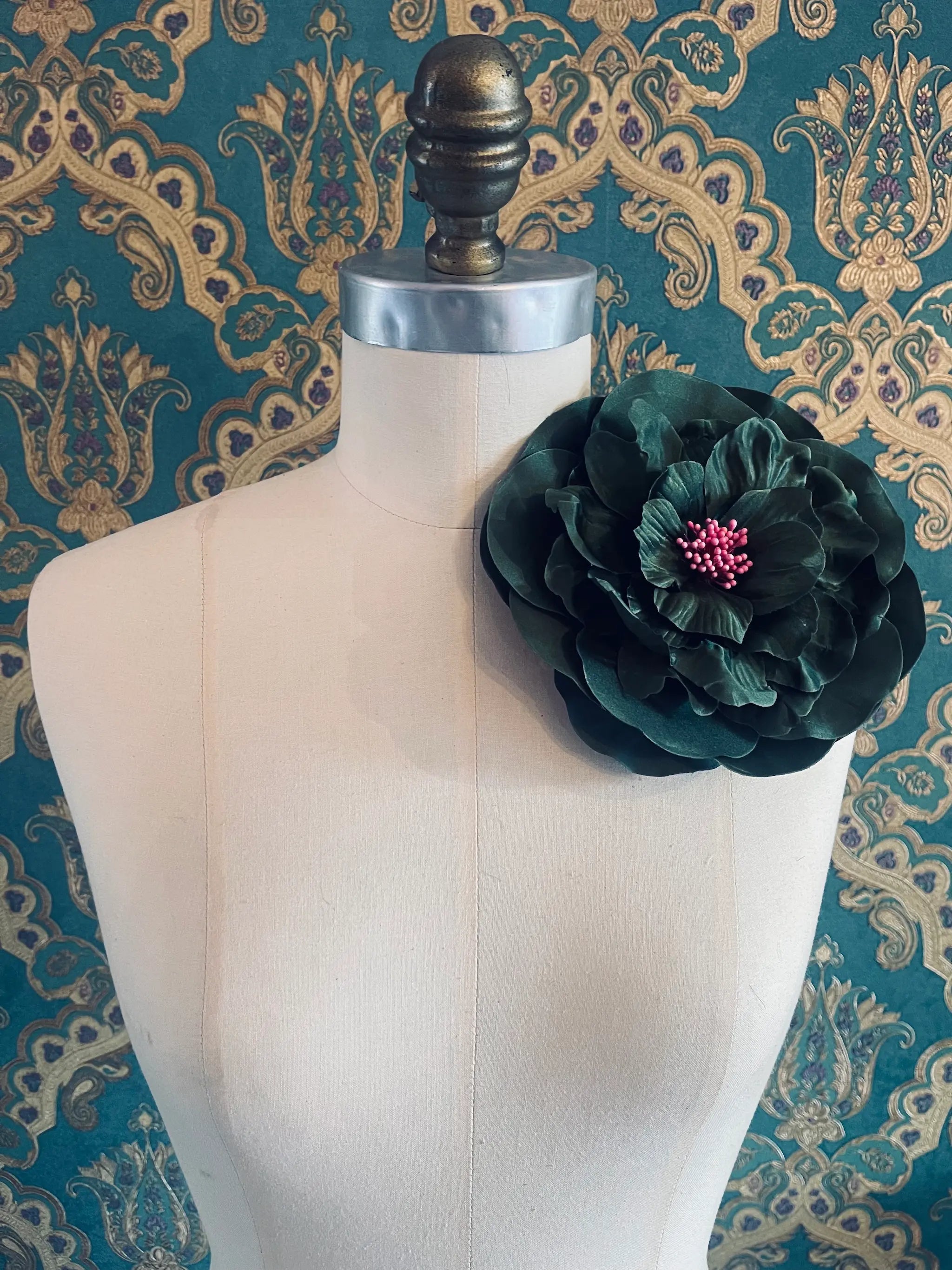 Broochella Ibisco large, green, handmade flower brooch. Styled on a mannequin. The background is green and gold damask wallpaper