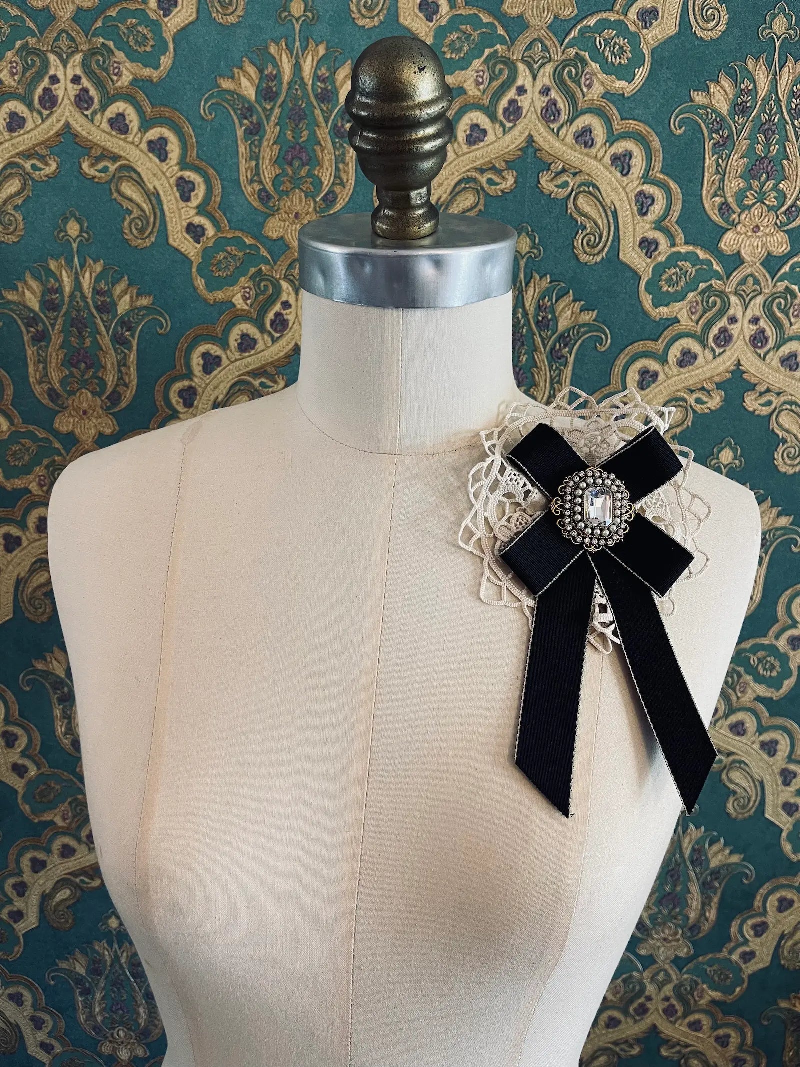 Broochella Il Conte black handmade bow brooch with rhinestone centre embellishment. Pictured on a mannequin. The background is green and gold damask wallpaper
