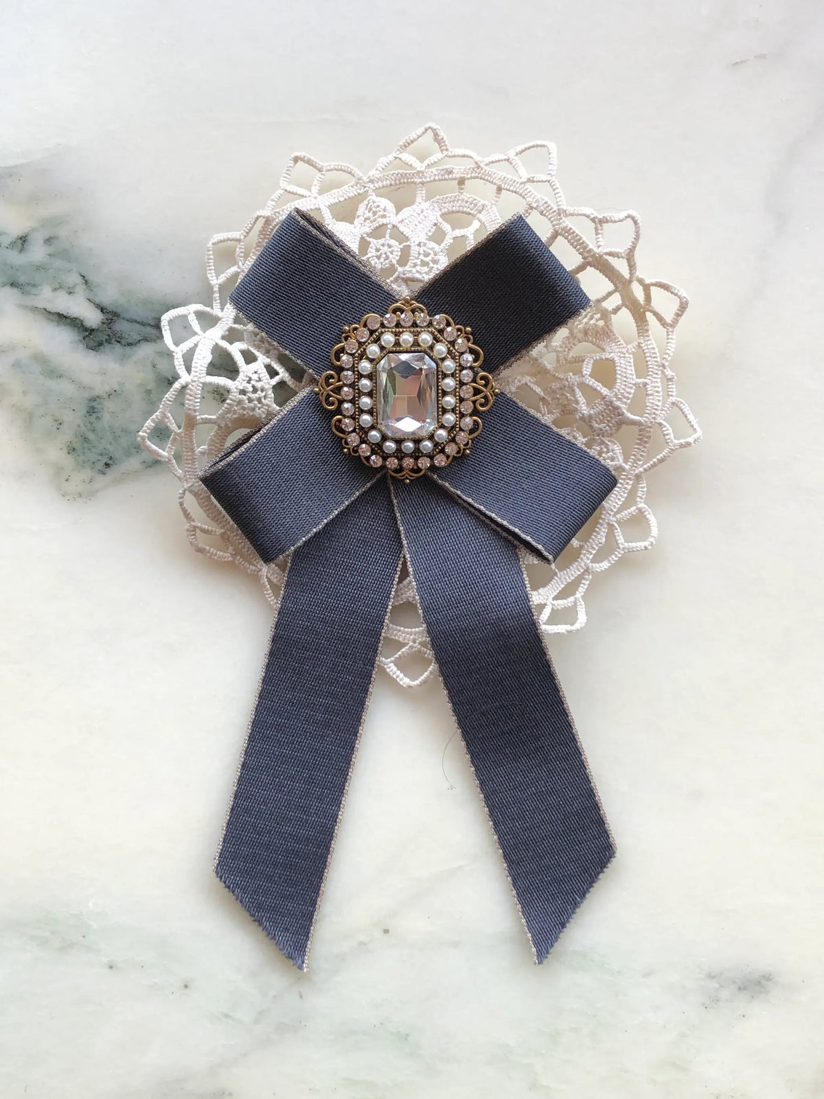 Broochella Il Conte Bow Brooch in grey ribbon with ivory lace and a pearl and crystal embellished centre, photographed on a white marble background with soft grey veining.