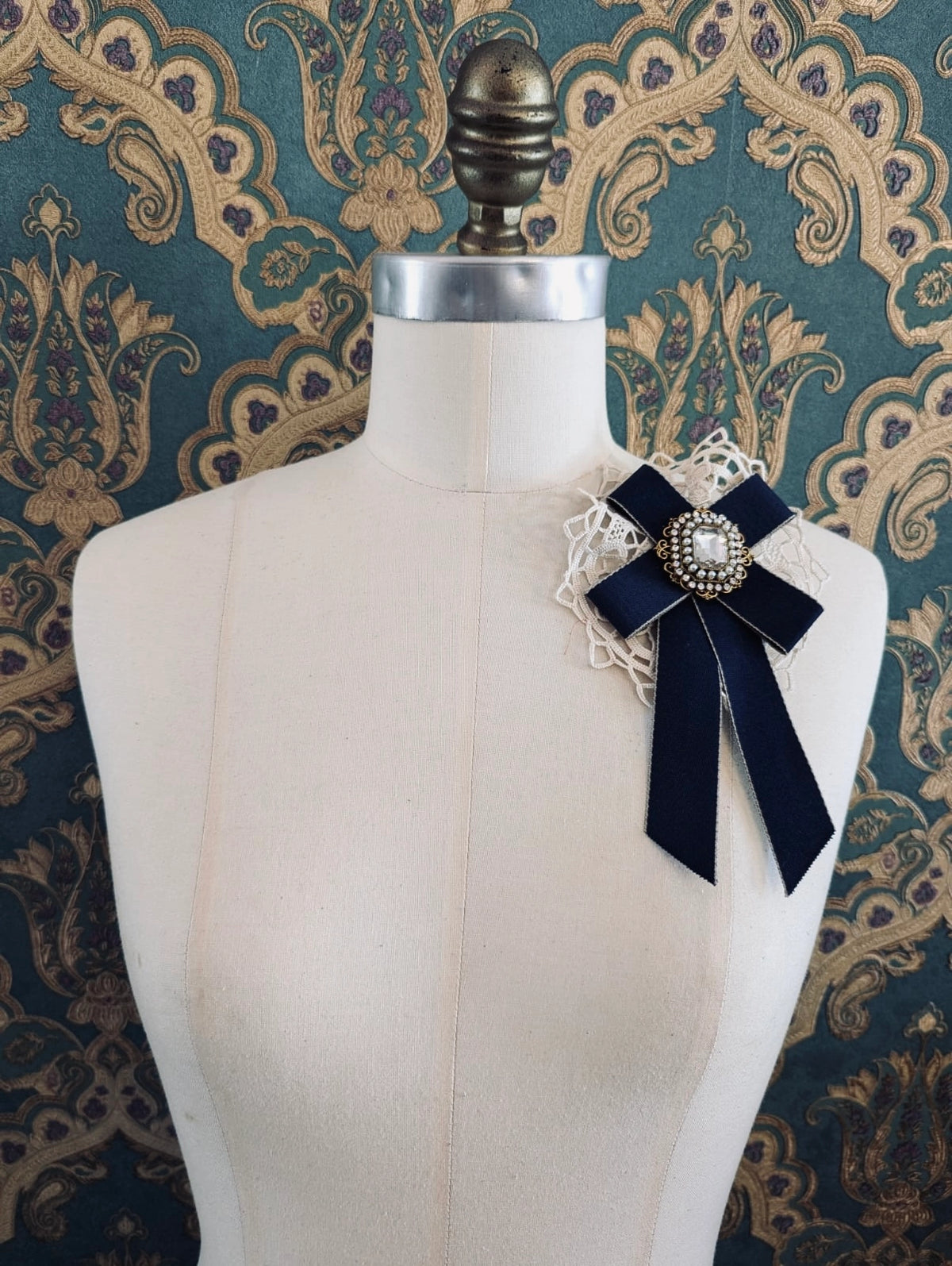 Broochella Il Conte navy handmade bow brooch with rhinestone centre embellishment. Pictured on a mannequin. The background is green and gold damask wallpaper