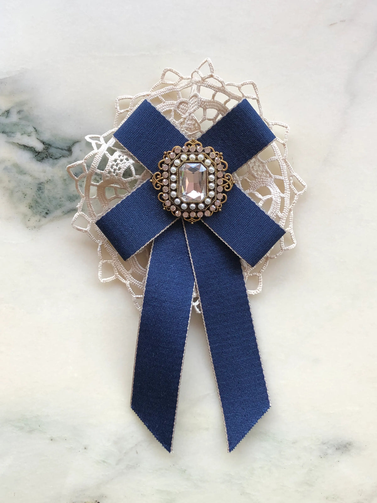 Broochella Il Conte Bow Brooch in navy ribbon with ivory lace and an ornate pearl and crystal embellished centre, photographed on a white marble background with soft grey veining.