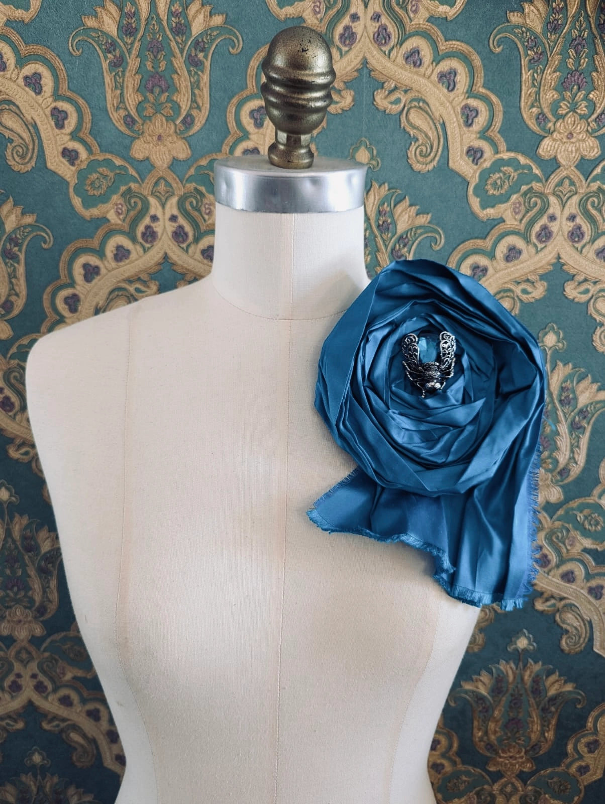 Broochella rosa ingioiellata large blue handmade silk flower brooch with an insect brooch embellishment in the centre. It is pinned to a mannequin. The background is green and gold damask wallpaper.