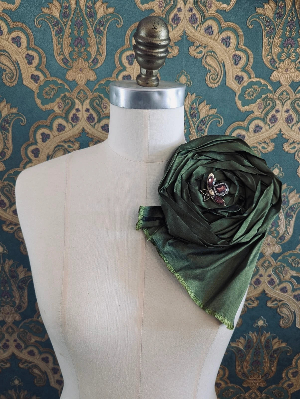 Broochella rosa ingioiellata large green handmade silk flower brooch with an insect brooch embellishment in the centre. It is pinned to a mannequin. The background is green and gold damask wallpaper.