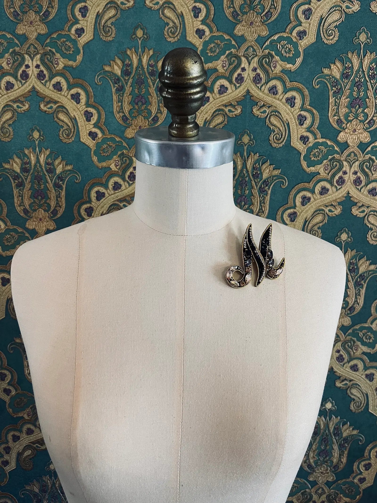 Broochella letter M hand beaded initial alphabet brooch in gold and black beads and sequins. Pictured on a mannequin. The background is green and gold damask wallpaper.