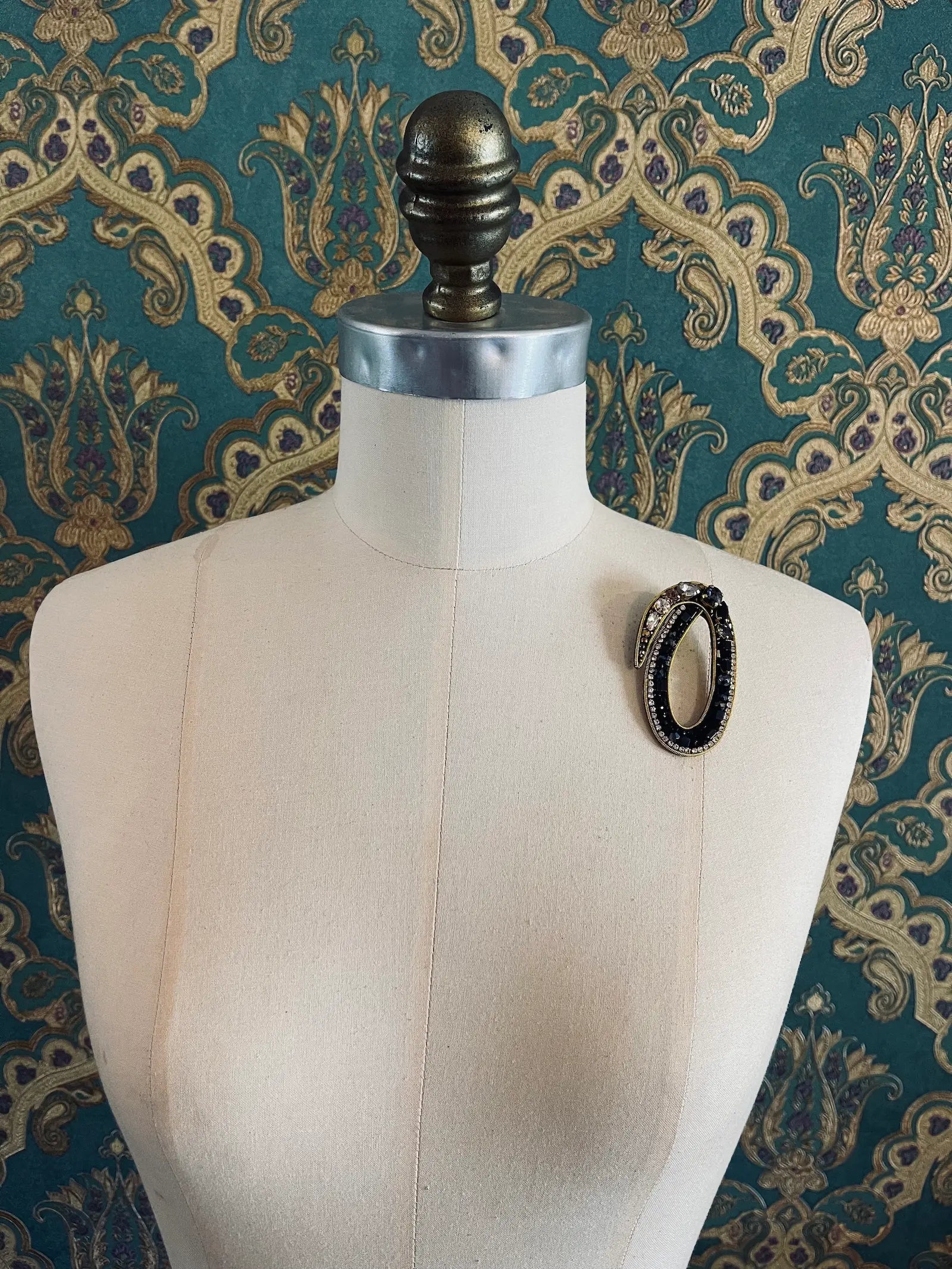 Broochella letter O hand beaded initial alphabet brooch in gold and black beads and sequins. Pictured on a mannequin. The background is green and gold damask wallpaper.