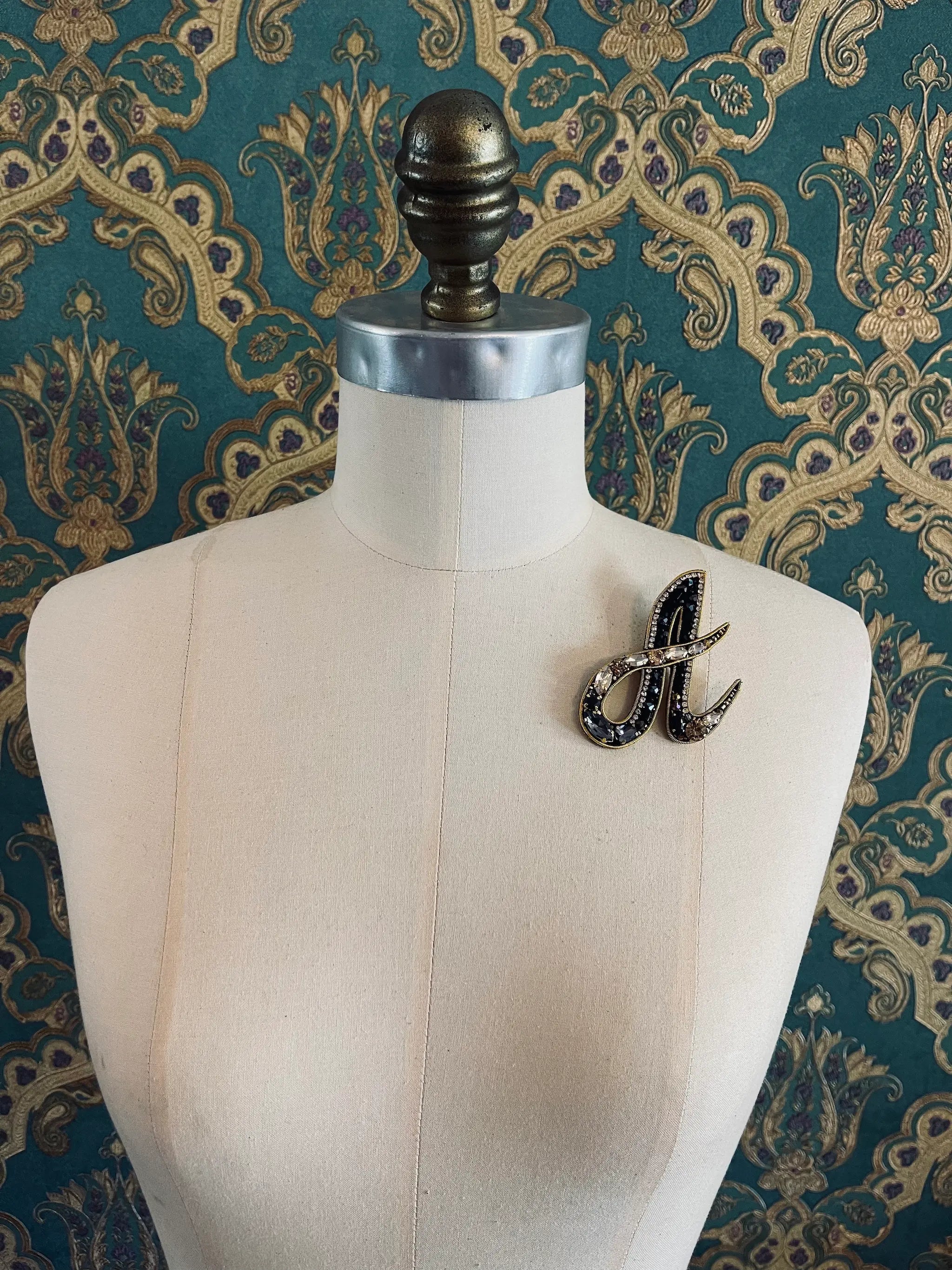 Broochella letter A hand beaded initial alphabet brooch in gold and black beads and sequins. Pictured on a mannequin. The background is green and gold damask wallpaper.