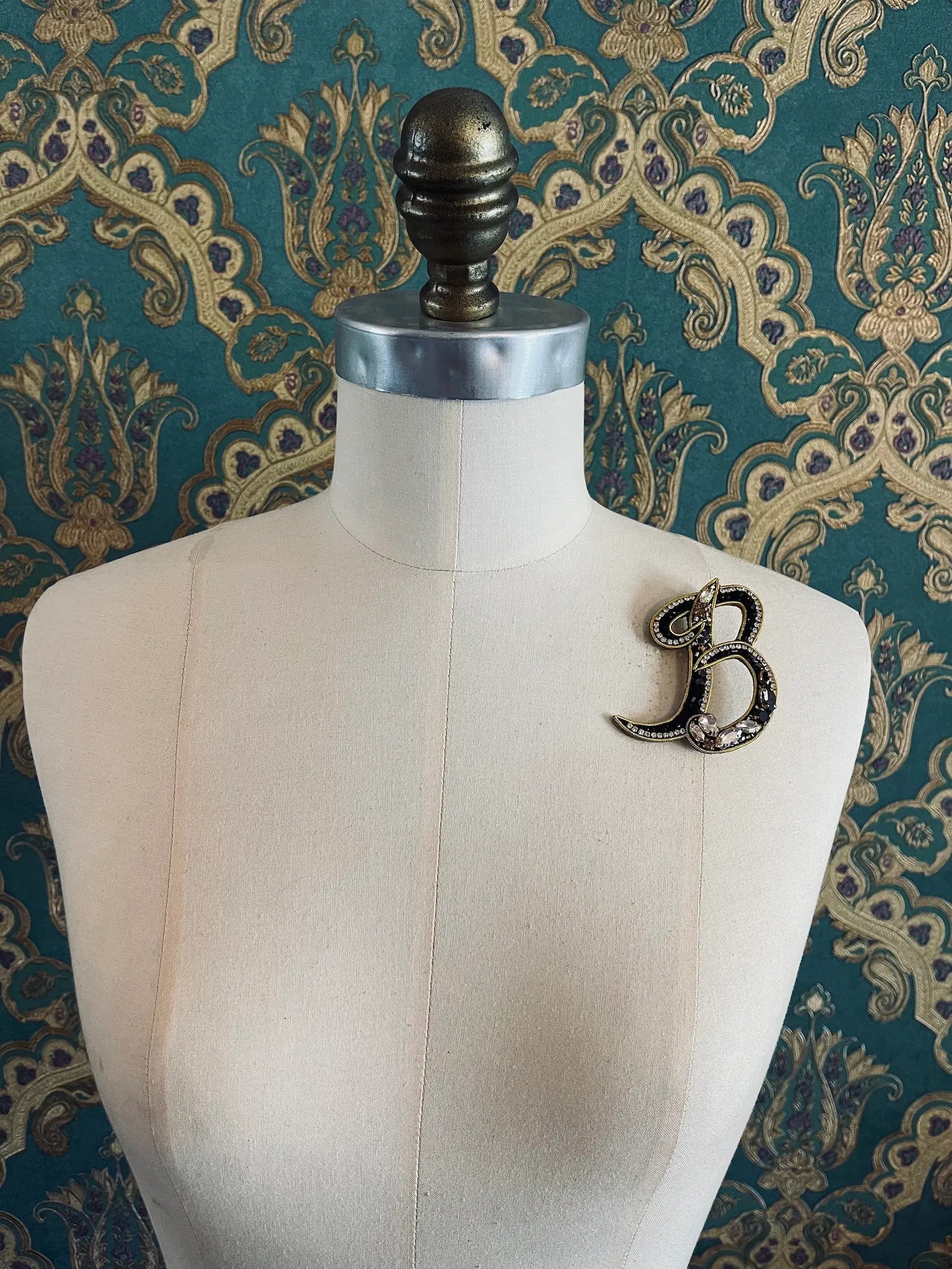 Broochella letter B hand beaded initial alphabet brooch in gold and black beads and sequins. Pictured on a mannequin. The background is green and gold damask wallpaper.