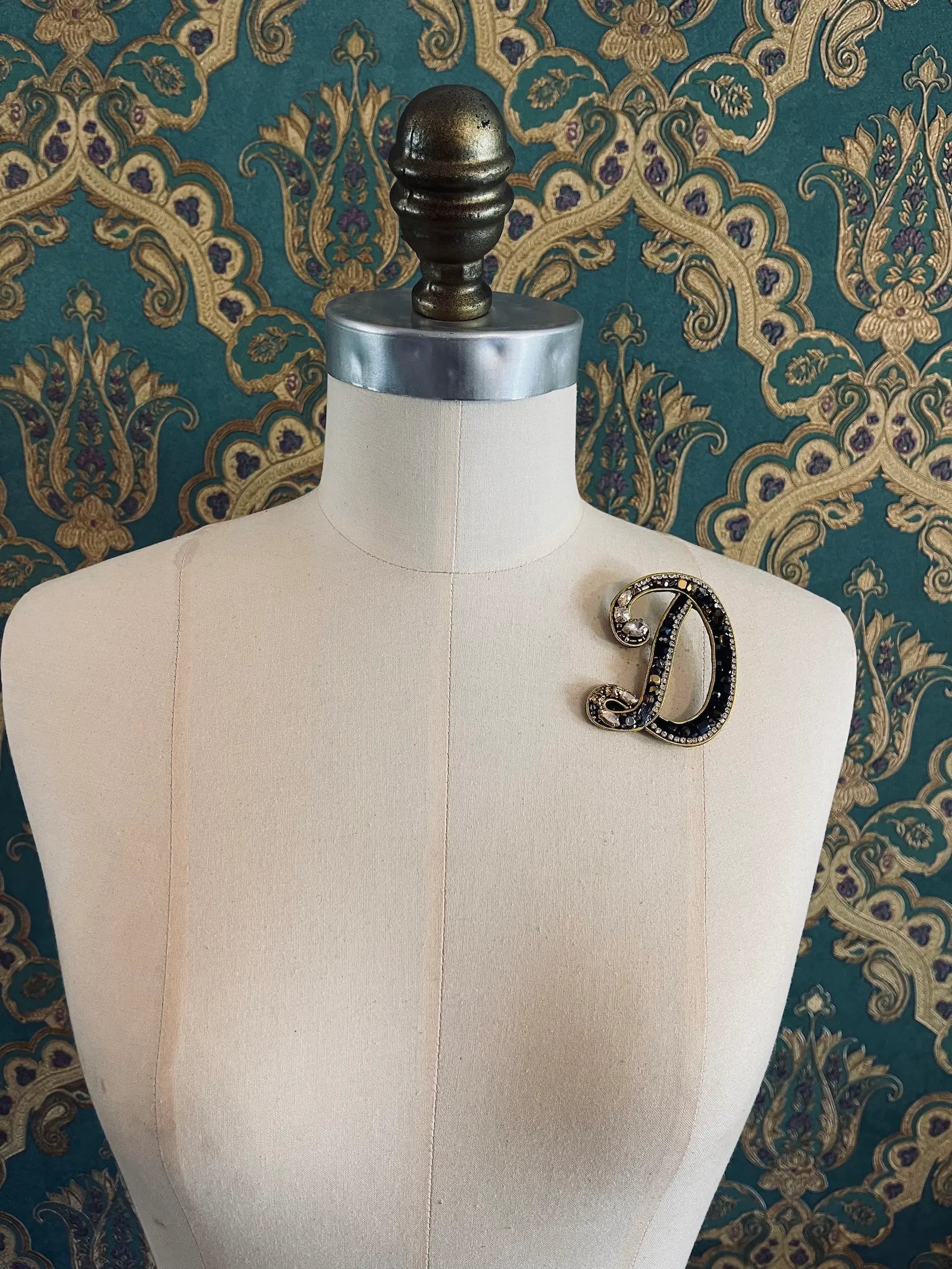 Broochella letter D hand beaded initial alphabet brooch in gold and black beads and sequins. Pictured on a mannequin. The background is green and gold damask wallpaper.