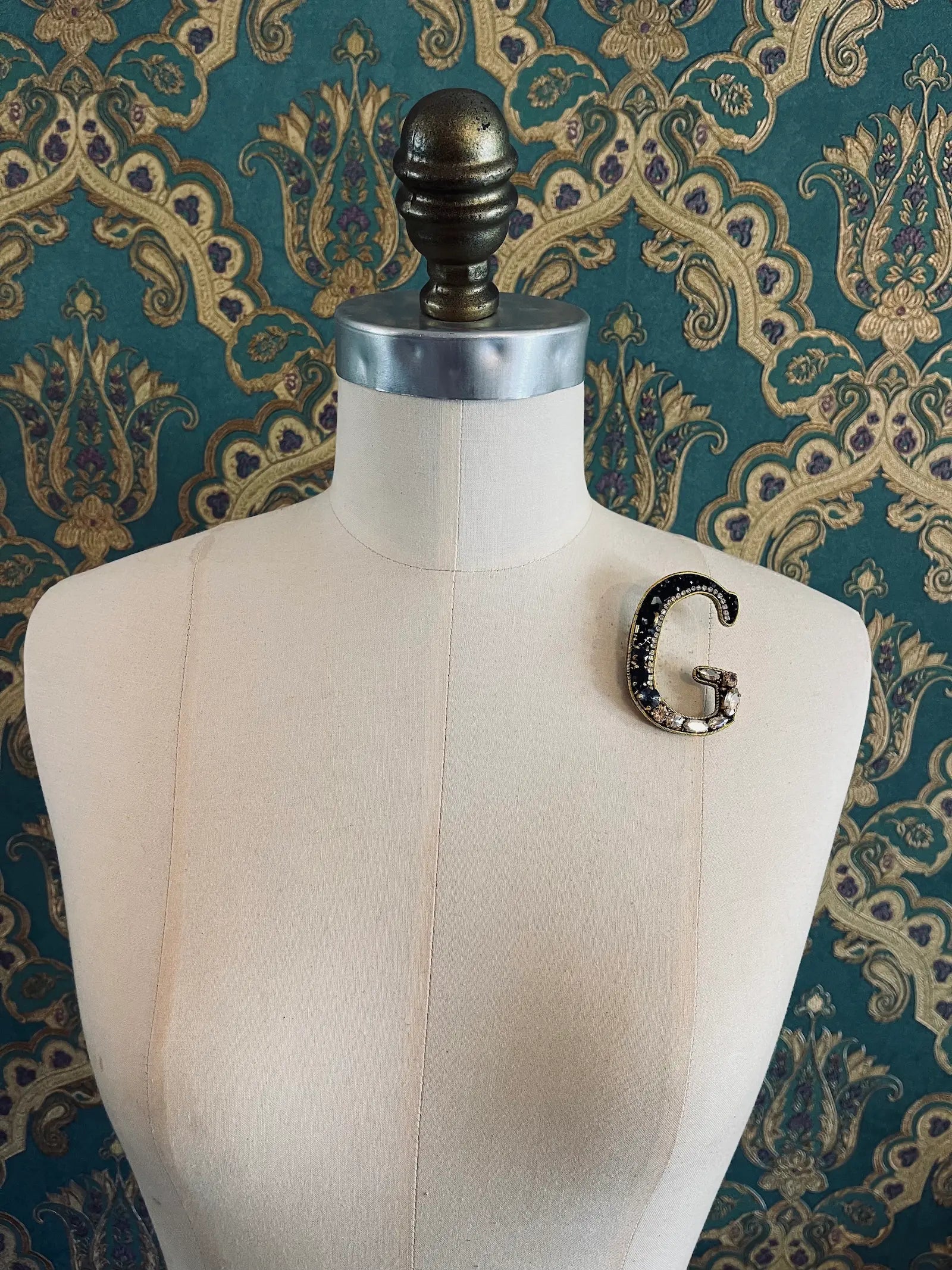 Broochella letter G hand beaded initial alphabet brooch in gold and black beads and sequins. Pictured on a mannequin. The background is green and gold damask wallpaper.
