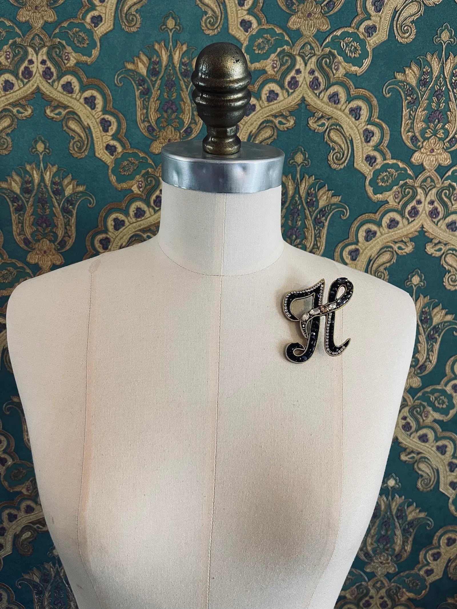 Broochella letter H hand beaded initial alphabet brooch in gold and black beads and sequins. Pictured on a mannequin. The background is green and gold damask wallpaper.
