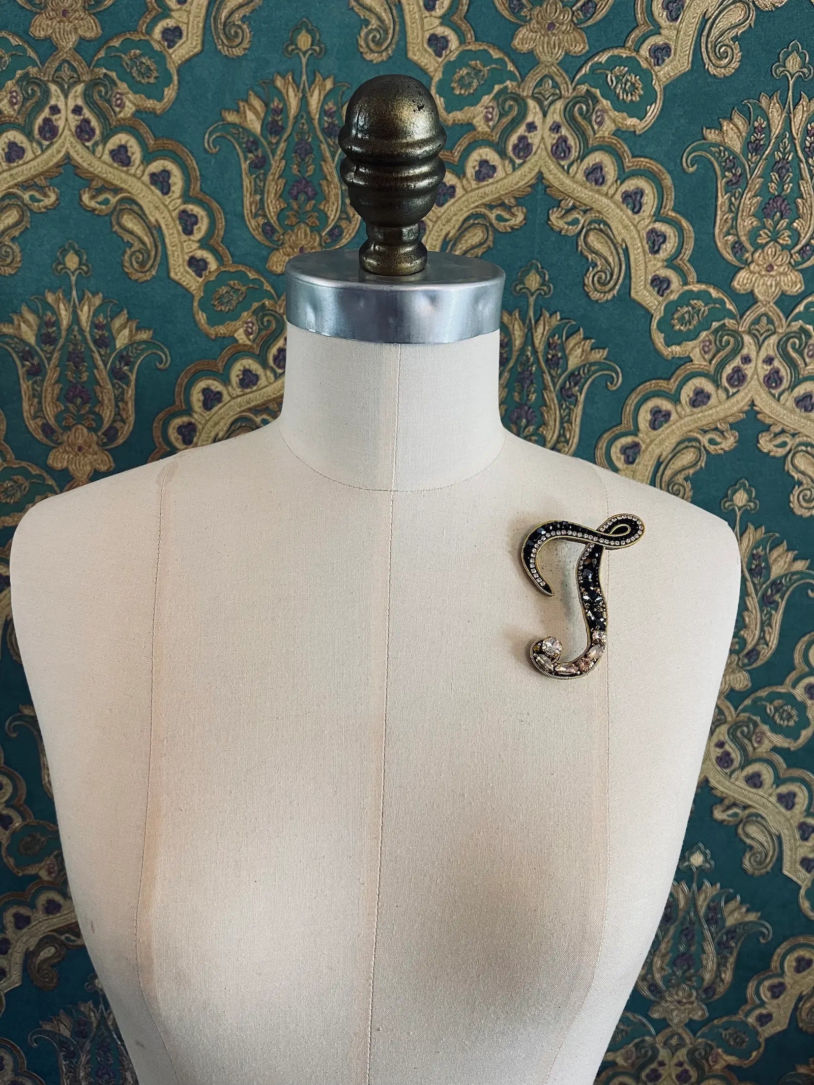 Broochella letter J hand beaded initial alphabet brooch in gold and black beads and sequins. Pictured on a mannequin. The background is green and gold damask wallpaper.