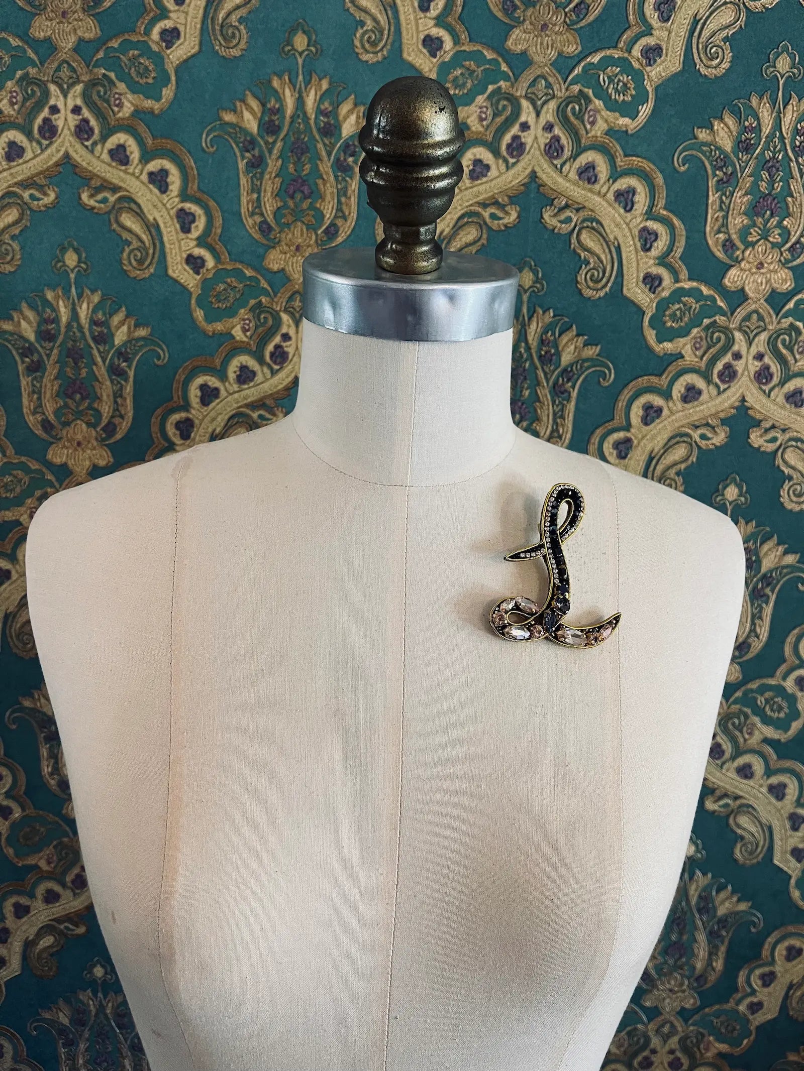 Broochella letter L hand beaded initial alphabet brooch in gold and black beads and sequins. Pictured on a mannequin. The background is green and gold damask wallpaper.
