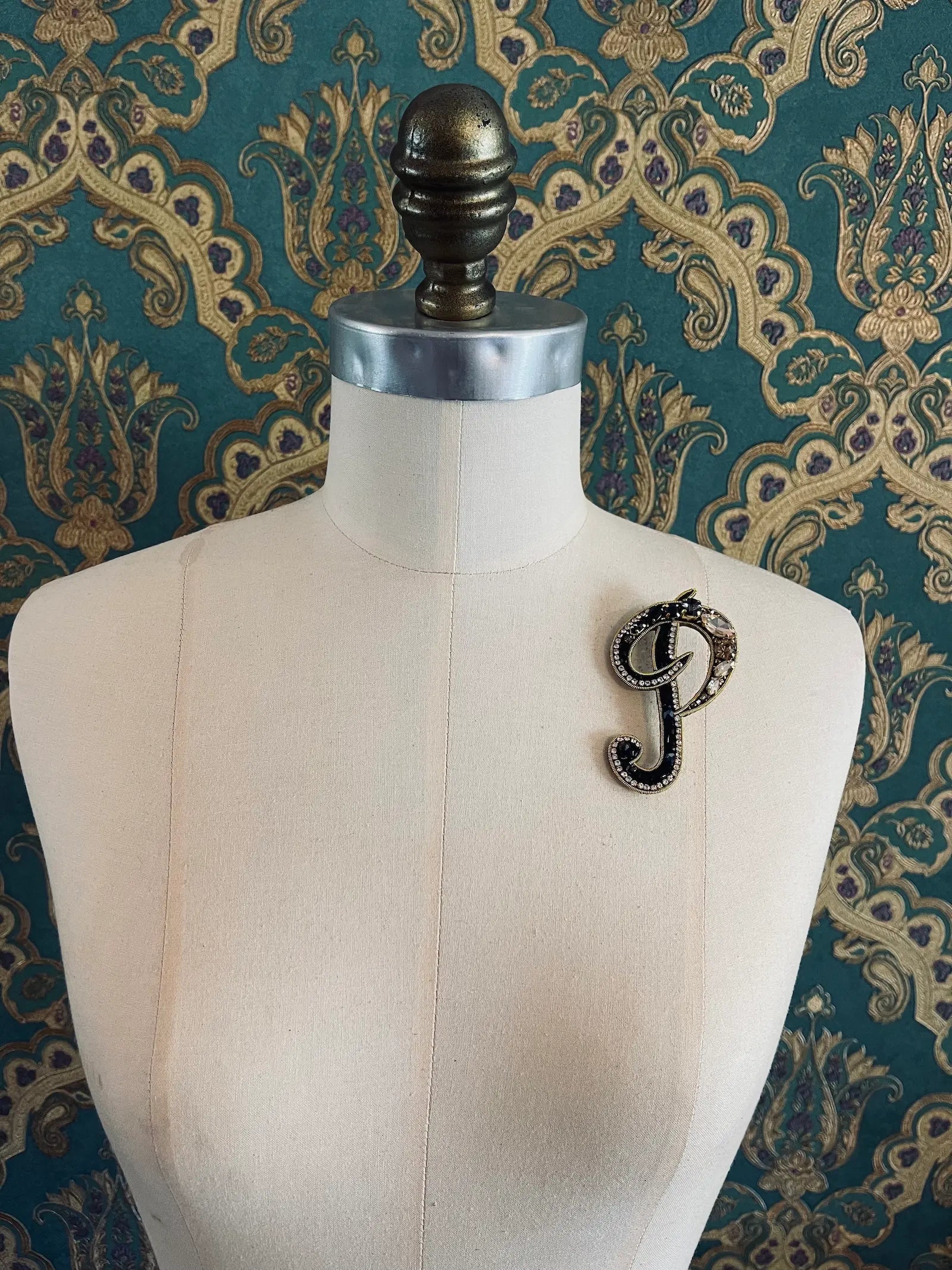 Broochella letter P hand beaded initial alphabet brooch in gold and black beads and sequins. Pictured on a mannequin. The background is green and gold damask wallpaper.