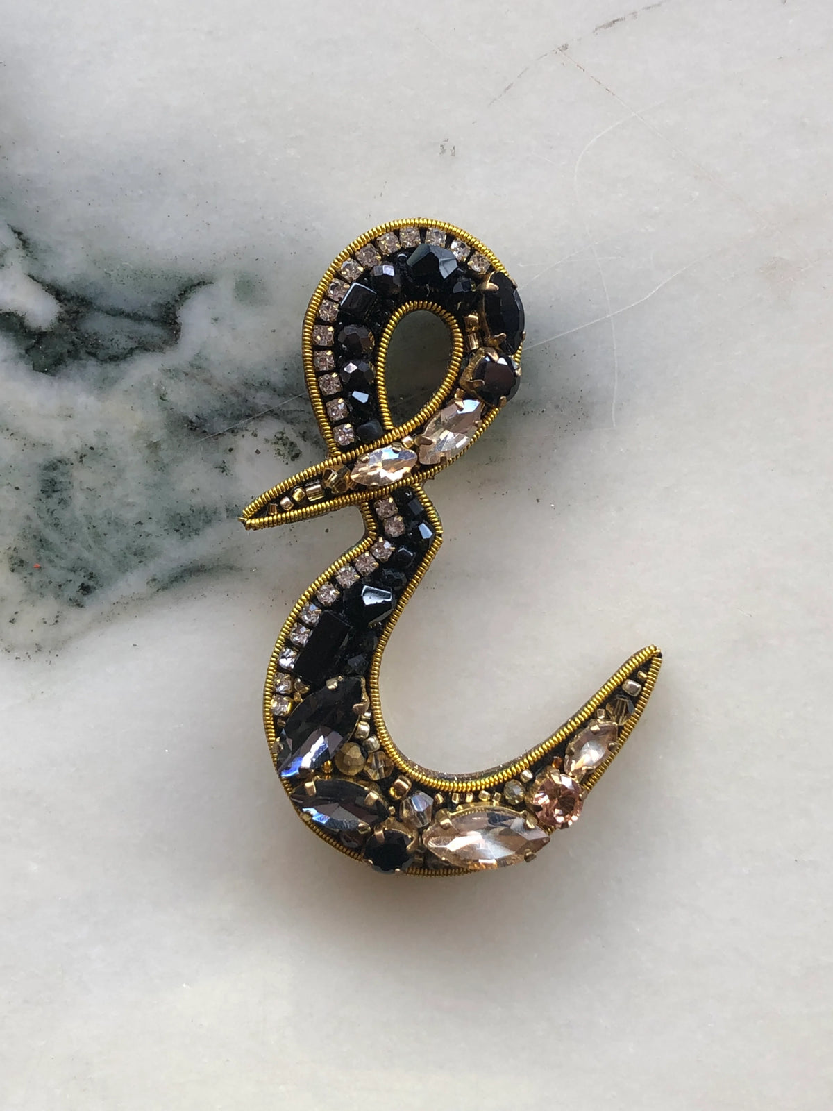 Broochella Initial E Brooch embellished with black beads, clear crystals and champagne stones with gold detailing, photographed on a white marble background with soft grey veining.