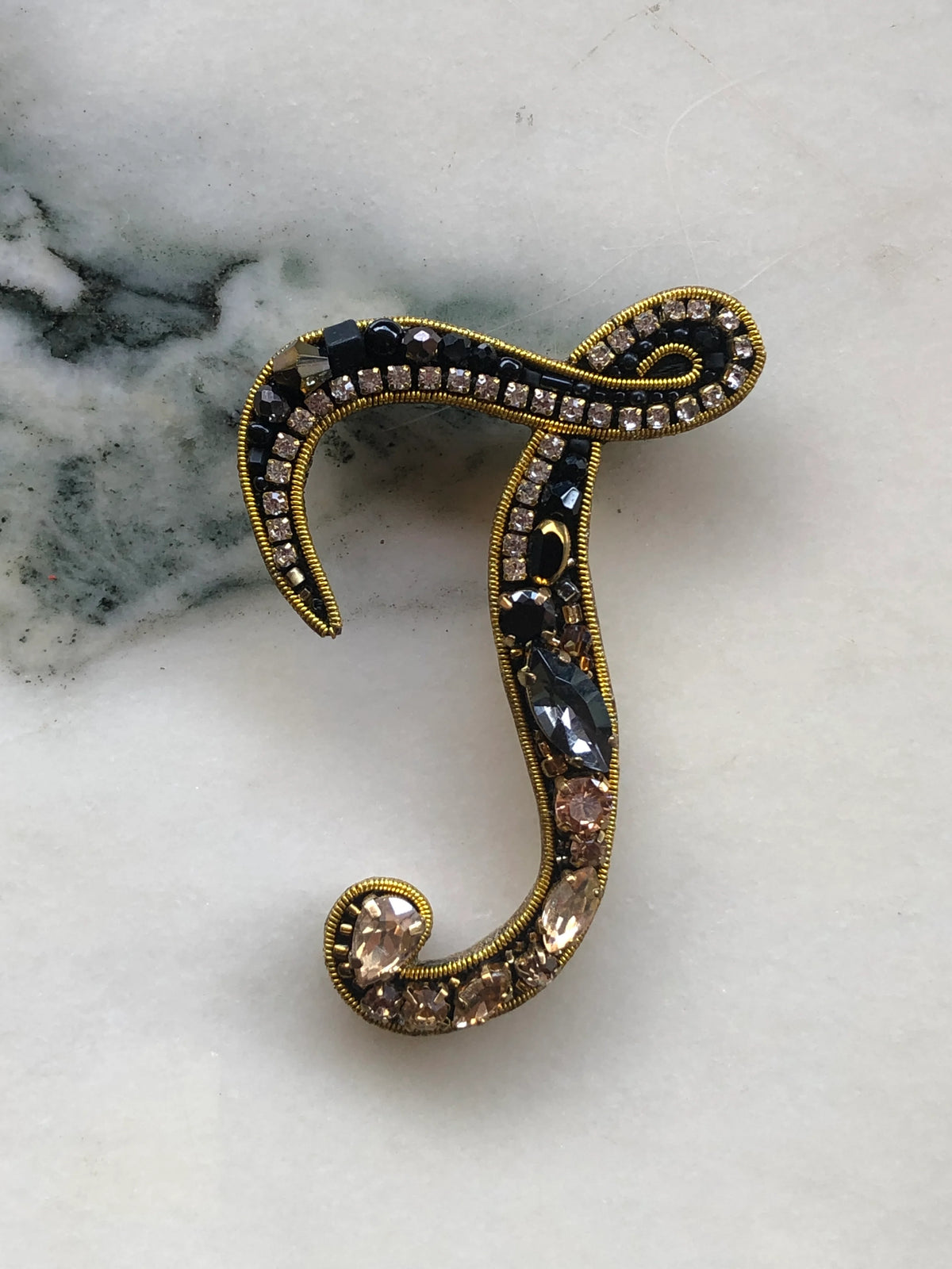 Broochella Initial J Brooch embellished with black beads, clear crystals and champagne stones with gold detailing, photographed on a white marble background with soft grey veining.