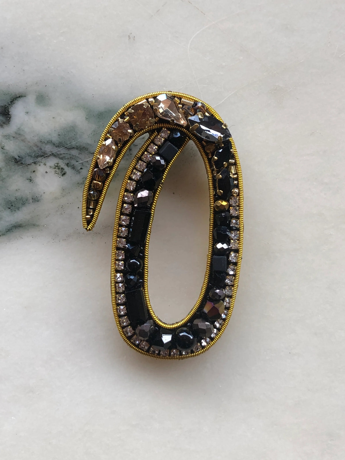 Broochella Initial O Brooch embellished with black beads, clear crystals and champagne stones with gold detailing, photographed on a white marble background with soft grey veining.