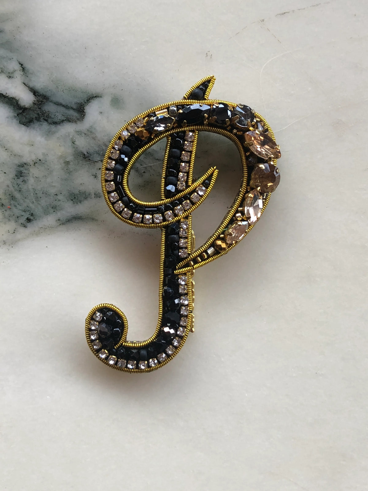 Broochella Initial P Brooch embellished with black beads, clear crystals and champagne stones with gold detailing, photographed on a white marble background with soft grey veining.