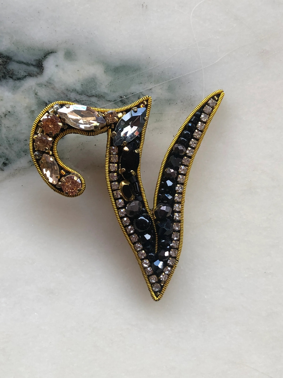 Broochella Initial V Brooch embellished with black beads, clear crystals and champagne stones with gold detailing, photographed on a white marble background with soft grey veining.