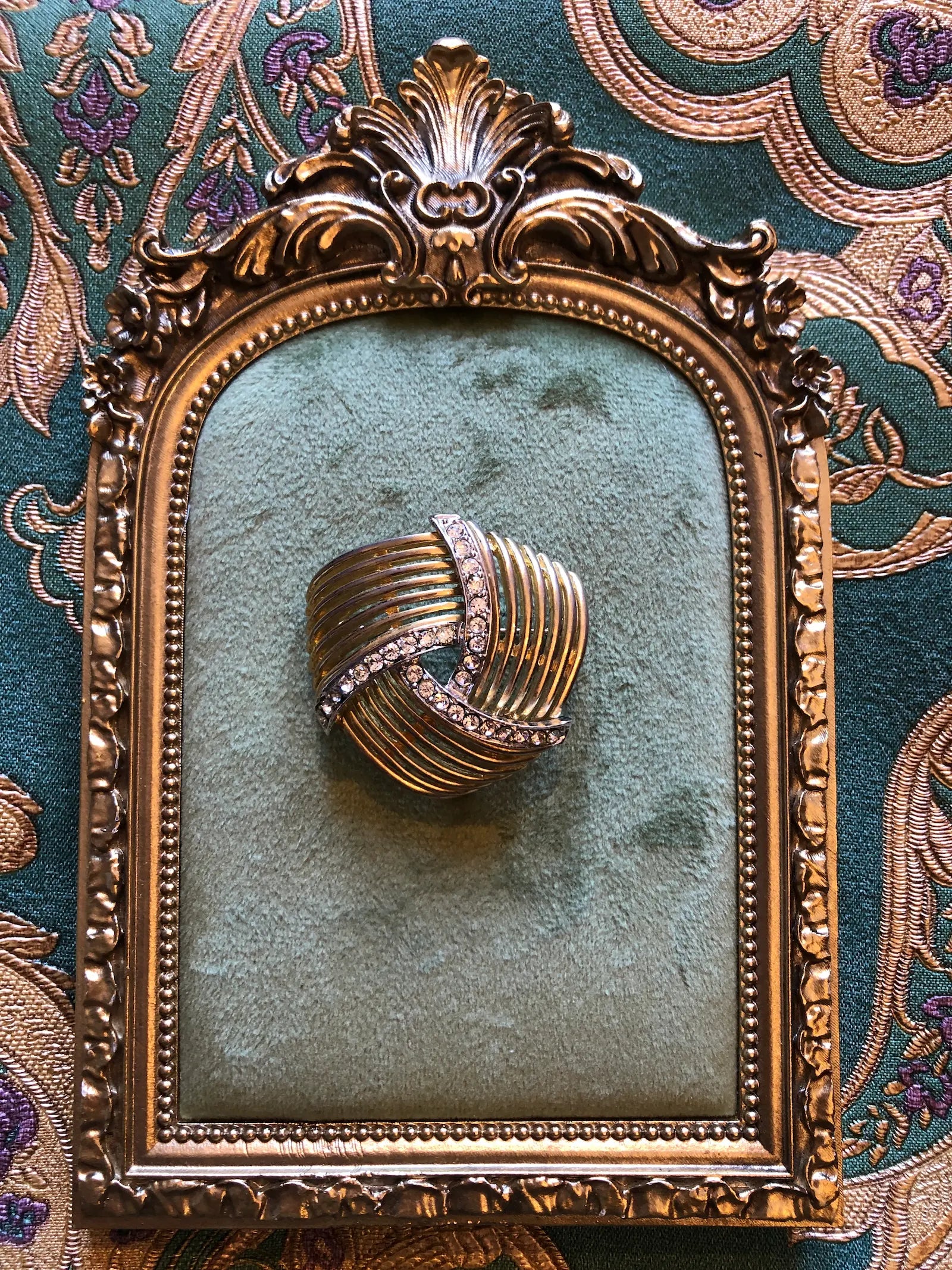 Broochella Intreccio vintage gold and silver-tone openwork brooch with sweeping ribbed bands and clear crystal accents. Pictured on a green velvet cushion inside an ornate gold picture frame against a patterned green and purple fabric background.