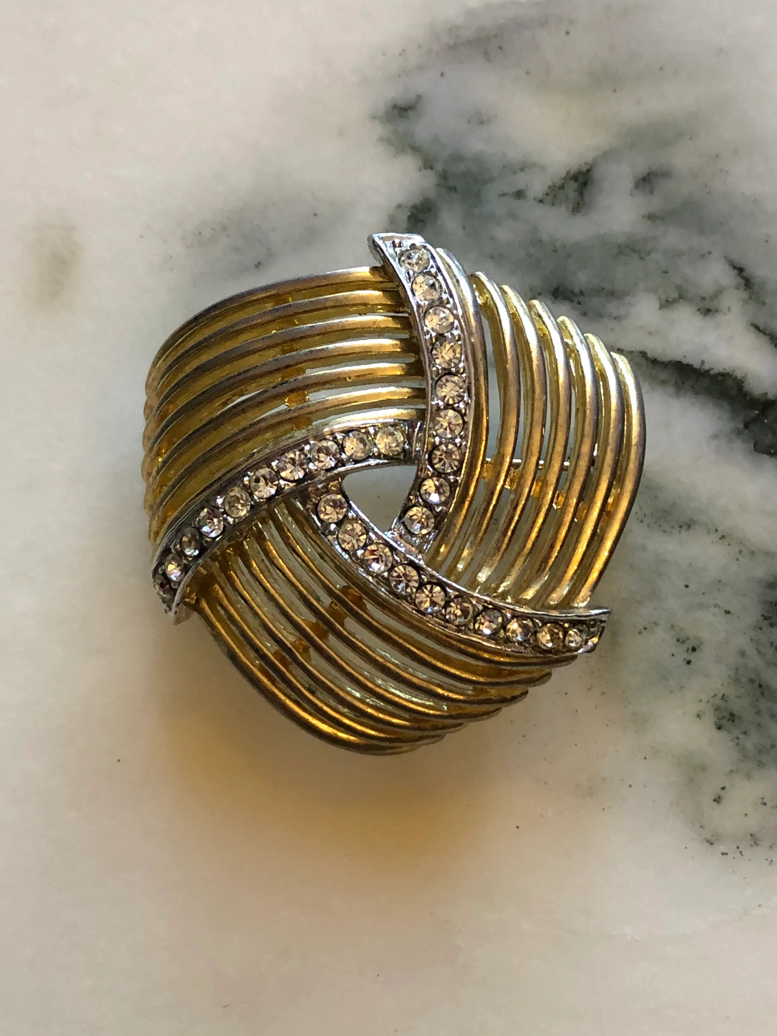 Broochella Intreccio vintage gold and silver-tone openwork brooch with sweeping ribbed bands and clear crystal accents. The background is white marble.