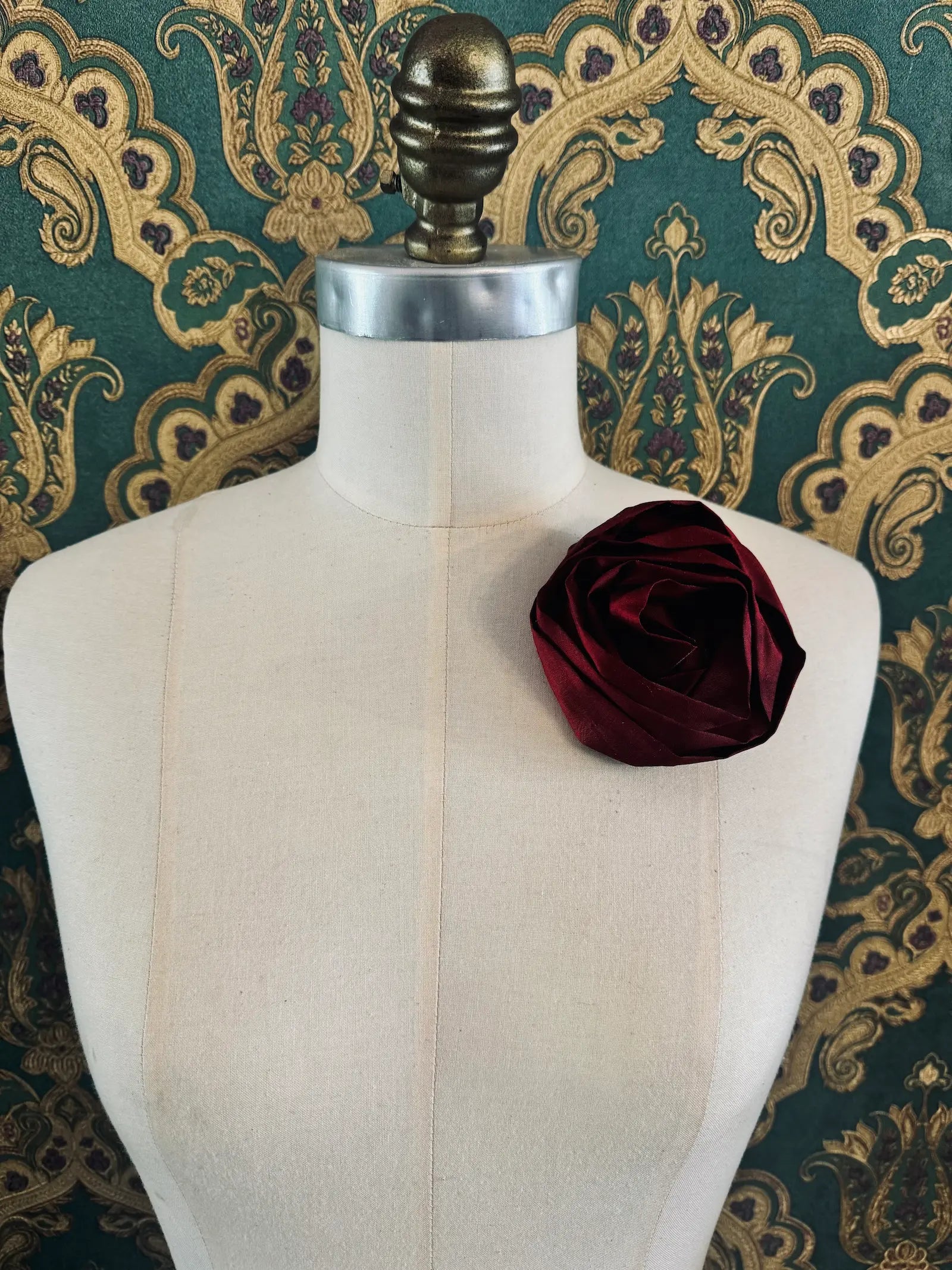Broochella Isabella Rossellini handmade silk rosette flower brooch in burgundy. Pictured on a mannequin.  The background is green and gold damask wallpaper