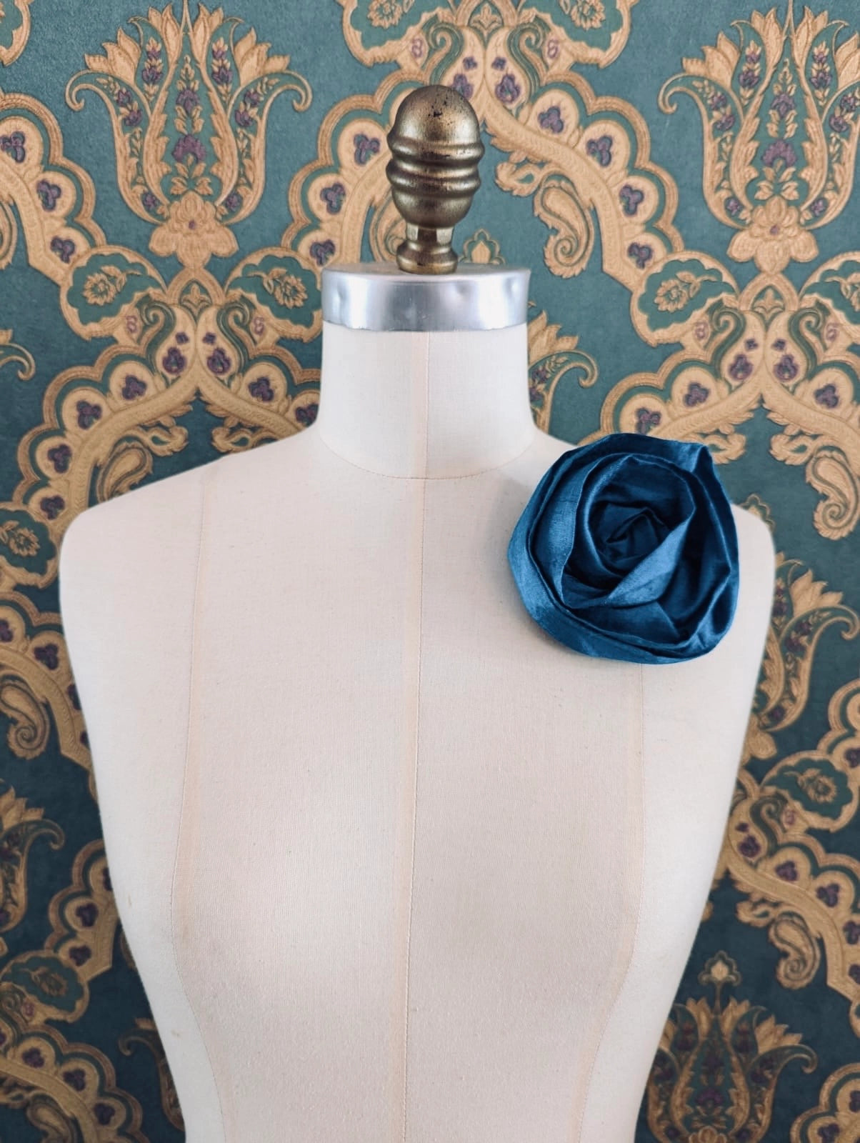 Broochella Isabella Rossellini handmade silk rosette flower brooch in peacock blue. Pictured on a mannequin.  The background is green and gold damask wallpaper