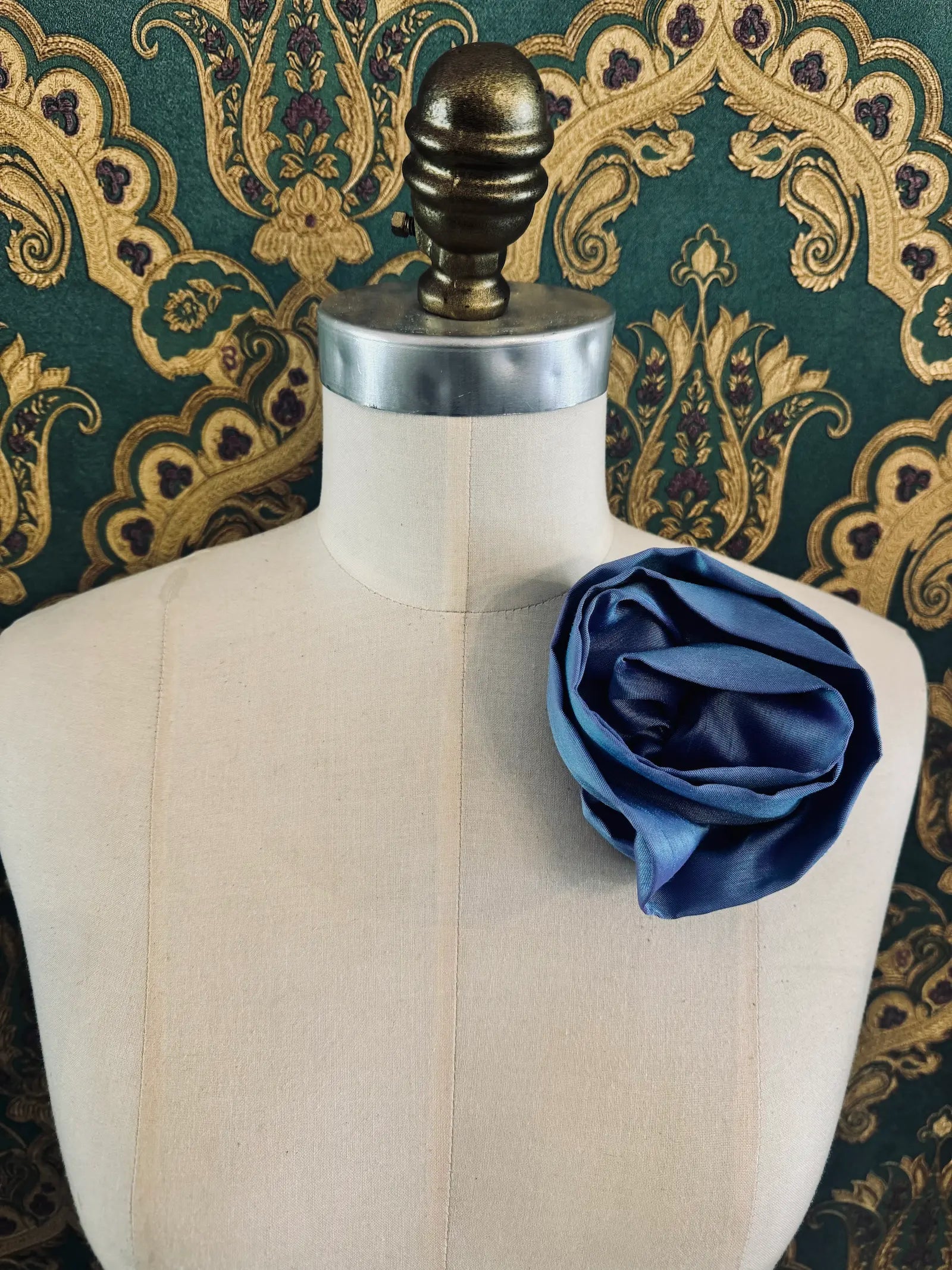 Broochella Isabella Rossellini handmade silk rosette flower brooch in powder blue. Pictured on a mannequin.  The background is green and gold damask wallpaper